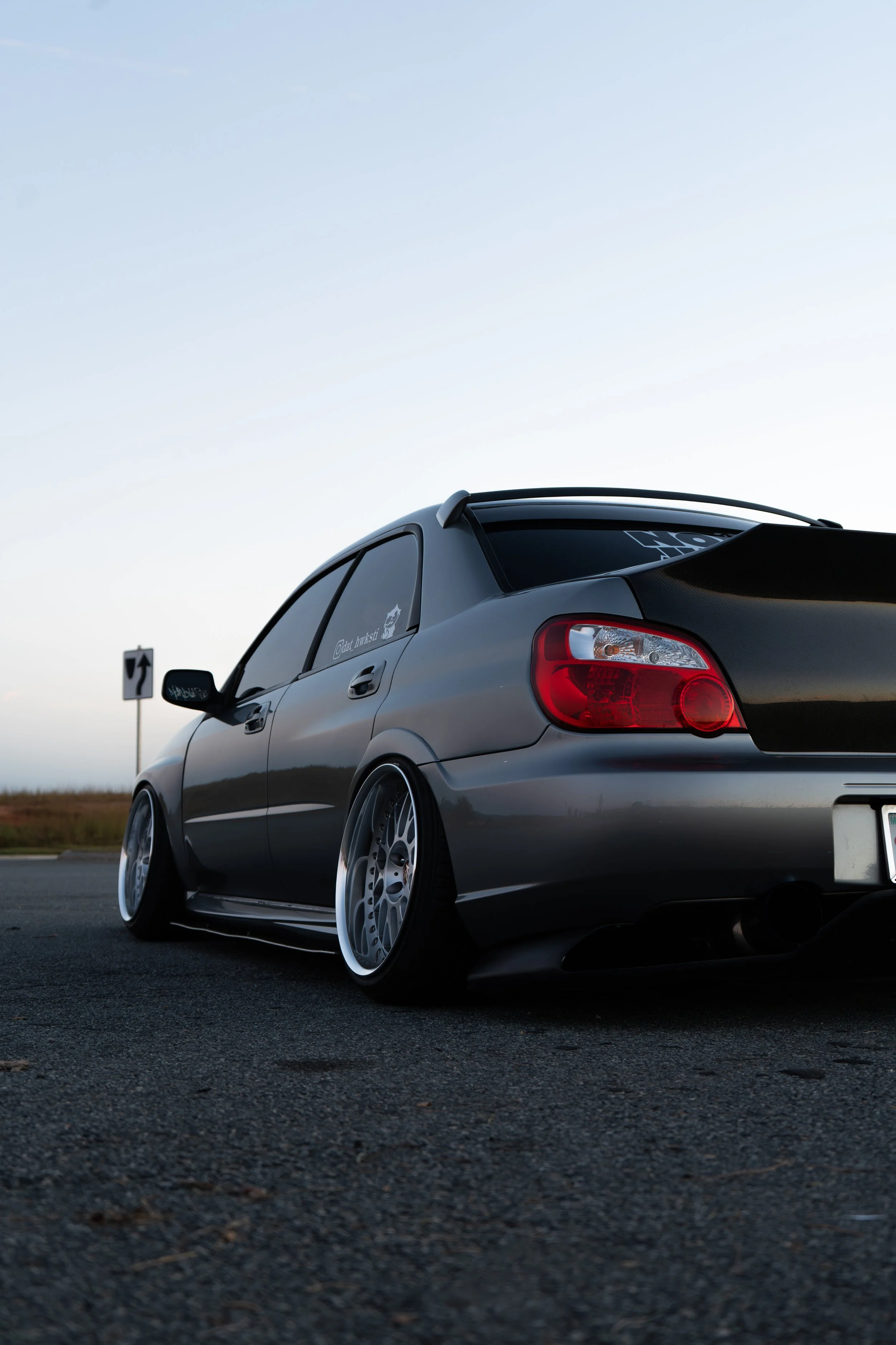 A lowered silver sports car with aftermarket wheels parked on an empty road during sunset.