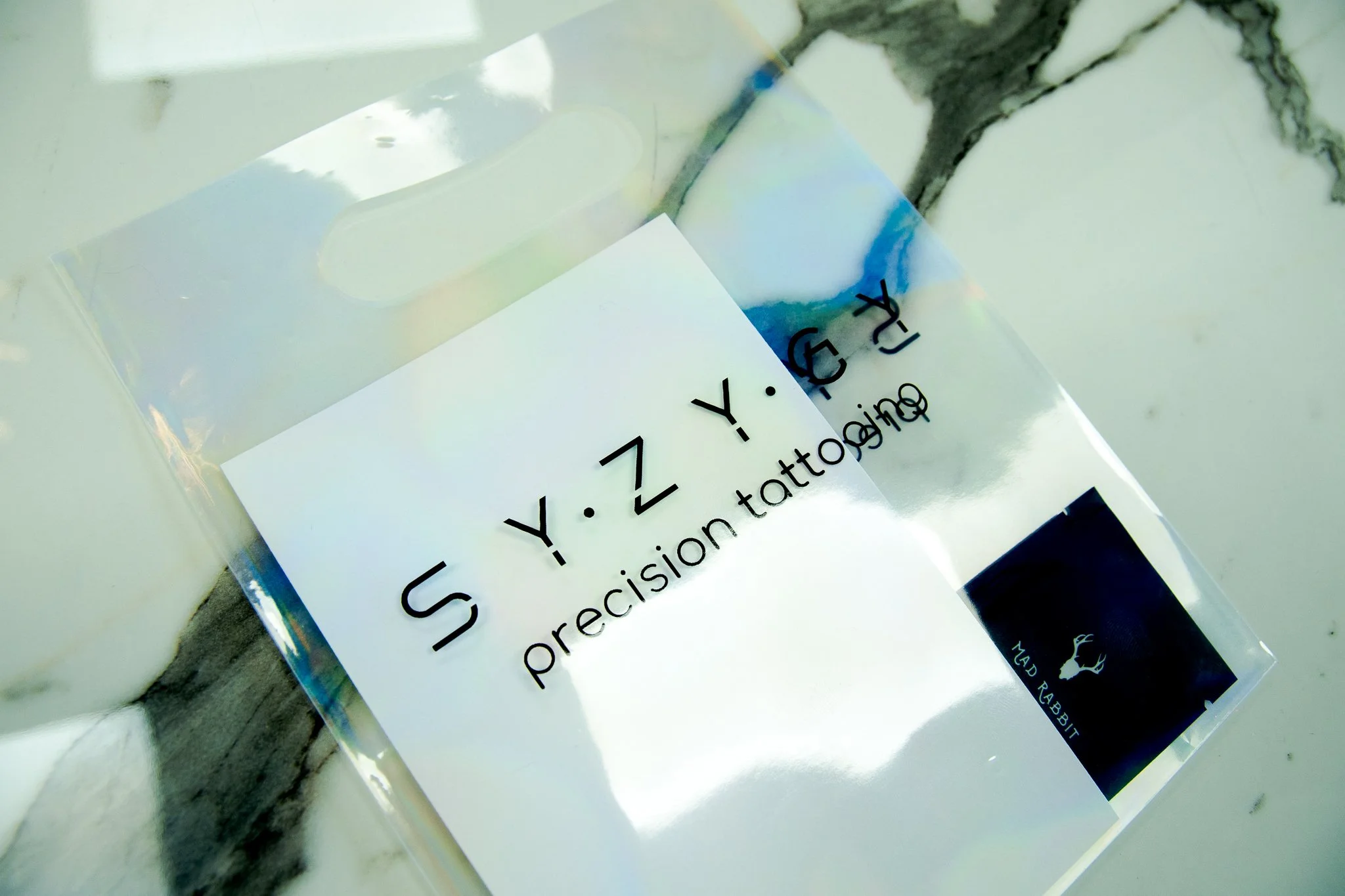 A plastic bag with a label reading 'S.Y.Z Y.G' and 'preciseion tattooing' on a marble surface, with a label from 'MAD RABBIT' in the corner.