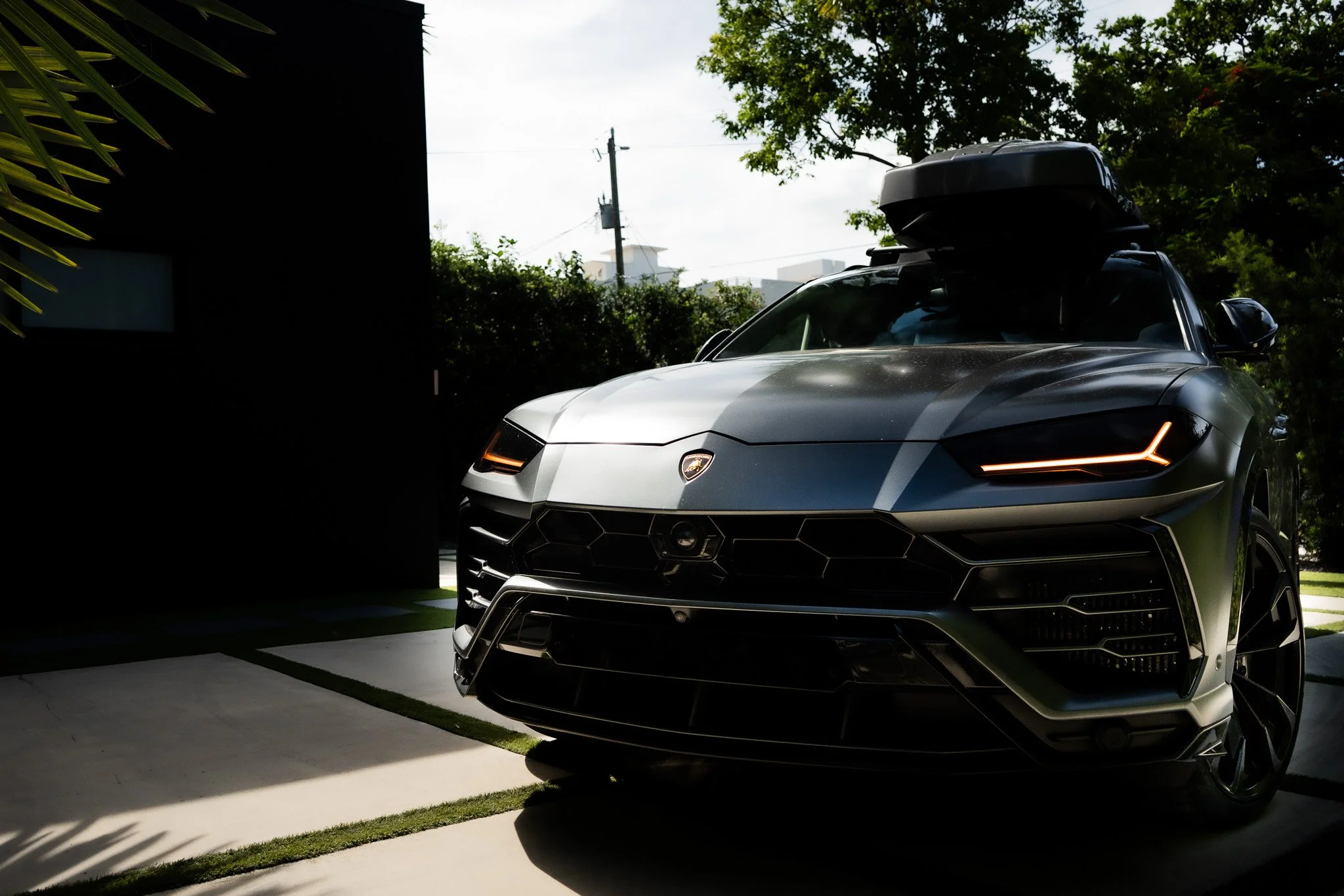 Black Lamborghini SUV with roof box parked on a driveway, with trees and cloudy sky in the background.