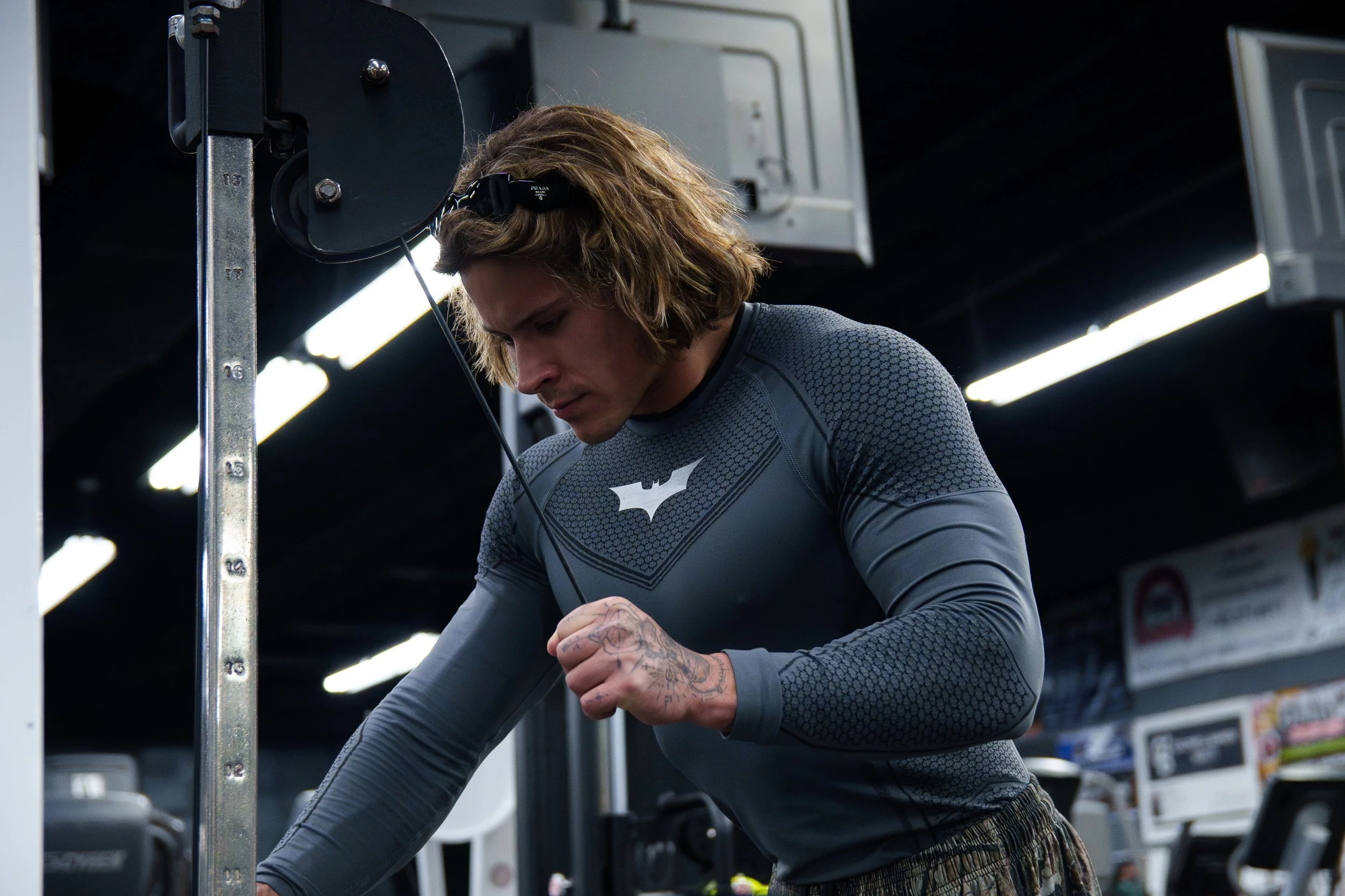 A young man with medium-length hair wearing a dark gray athletic shirt with a logo on the chest, in a gym, looking down while working out.