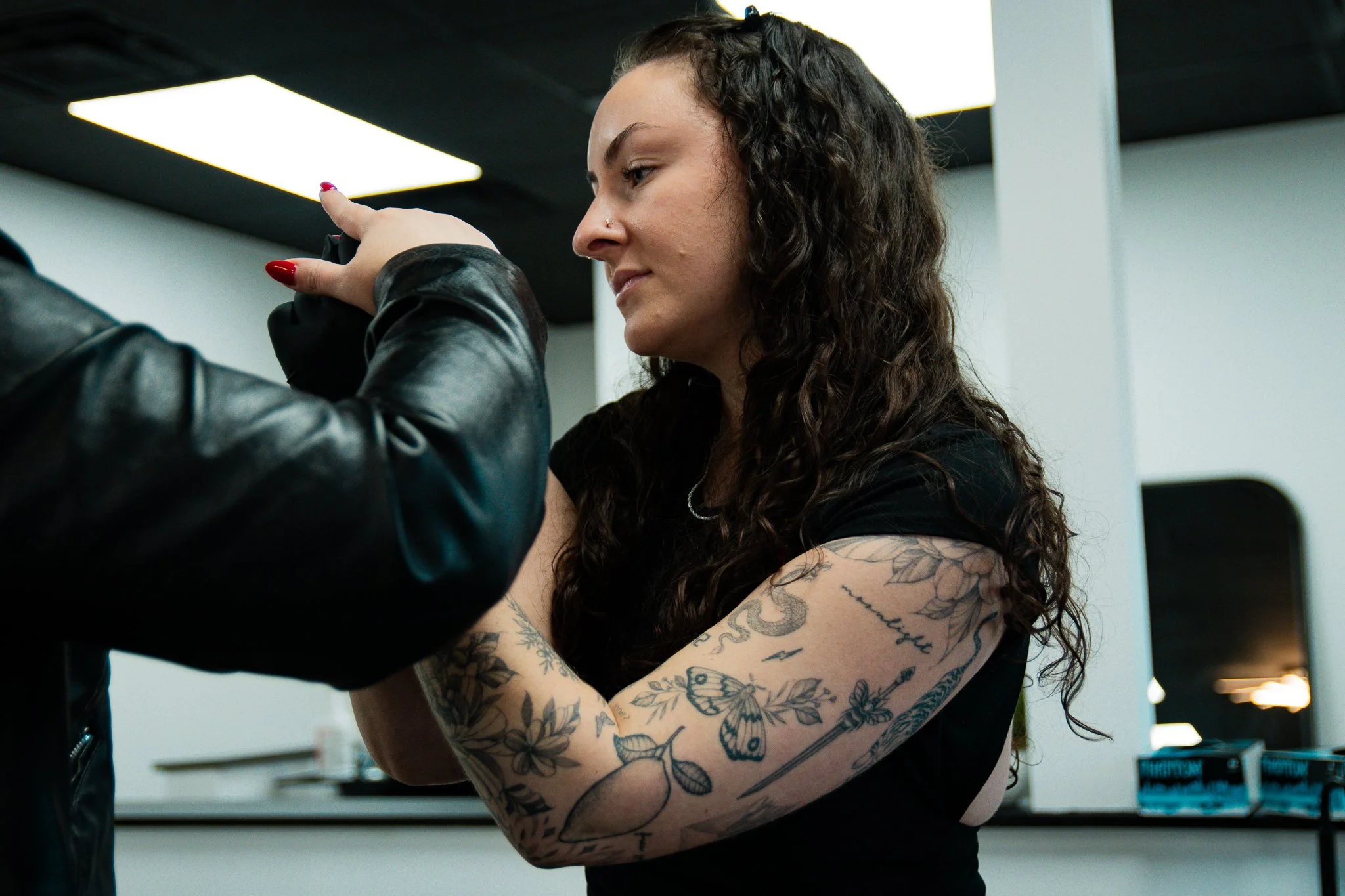 A woman with long, curly hair and tattoos on her arms getting a tattoo on her upper arm from an artist wearing a black leather jacket.