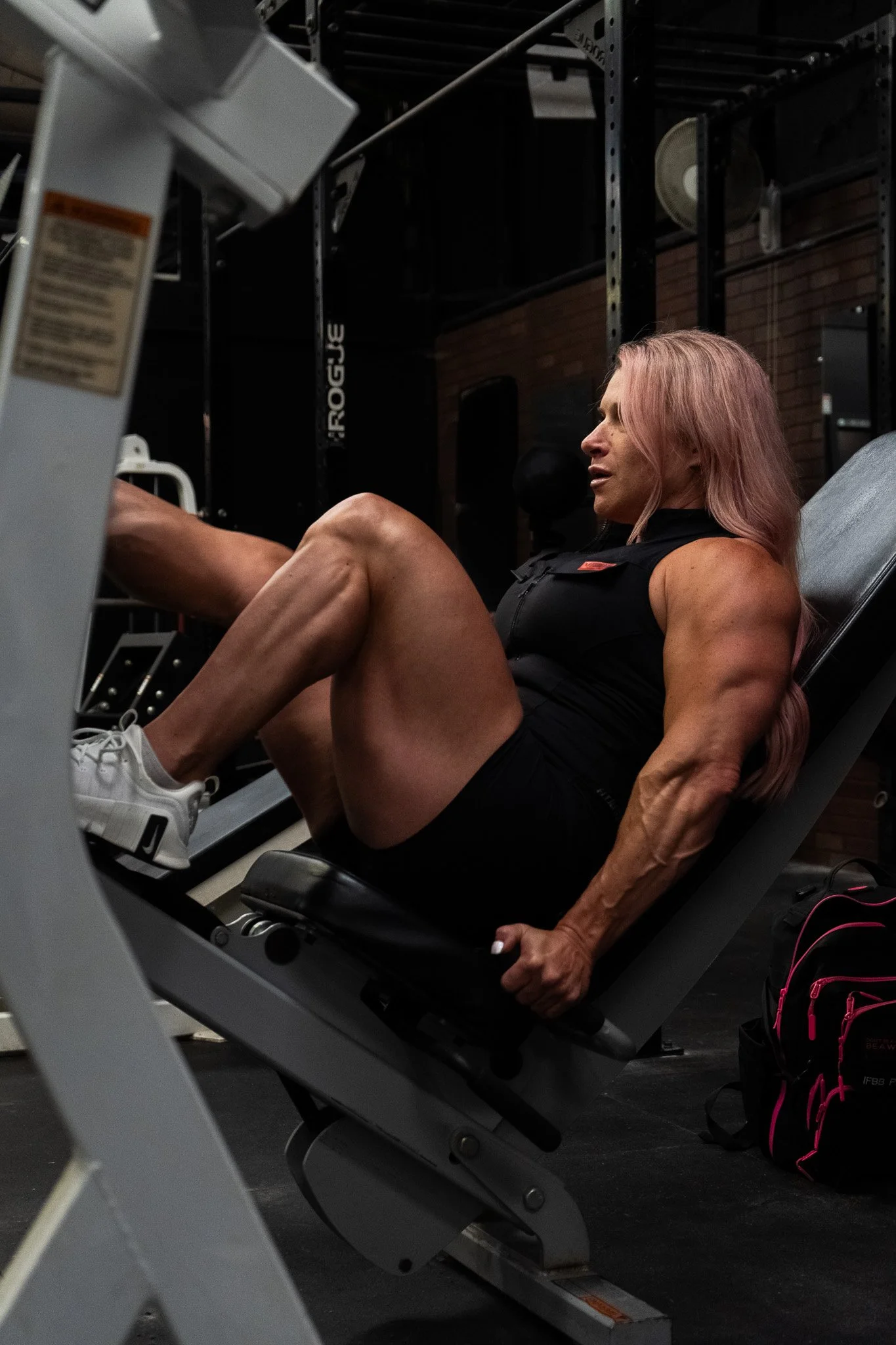 A muscular woman with long pink hair exercises on a leg press machine at the gym.