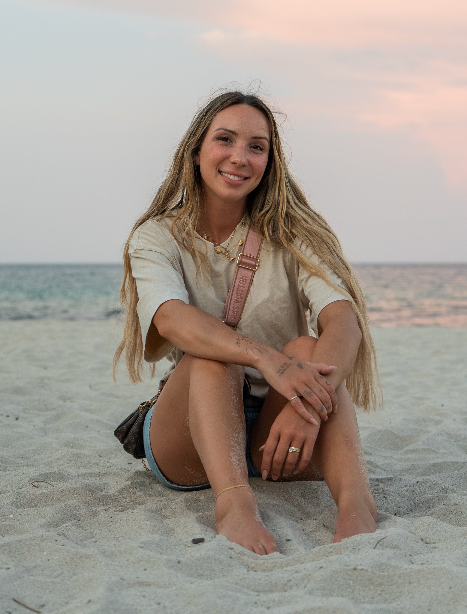 Young woman sitting on a beach at sunset with blond hair, wearing a beige t-shirt and shorts, smiling at the camera.