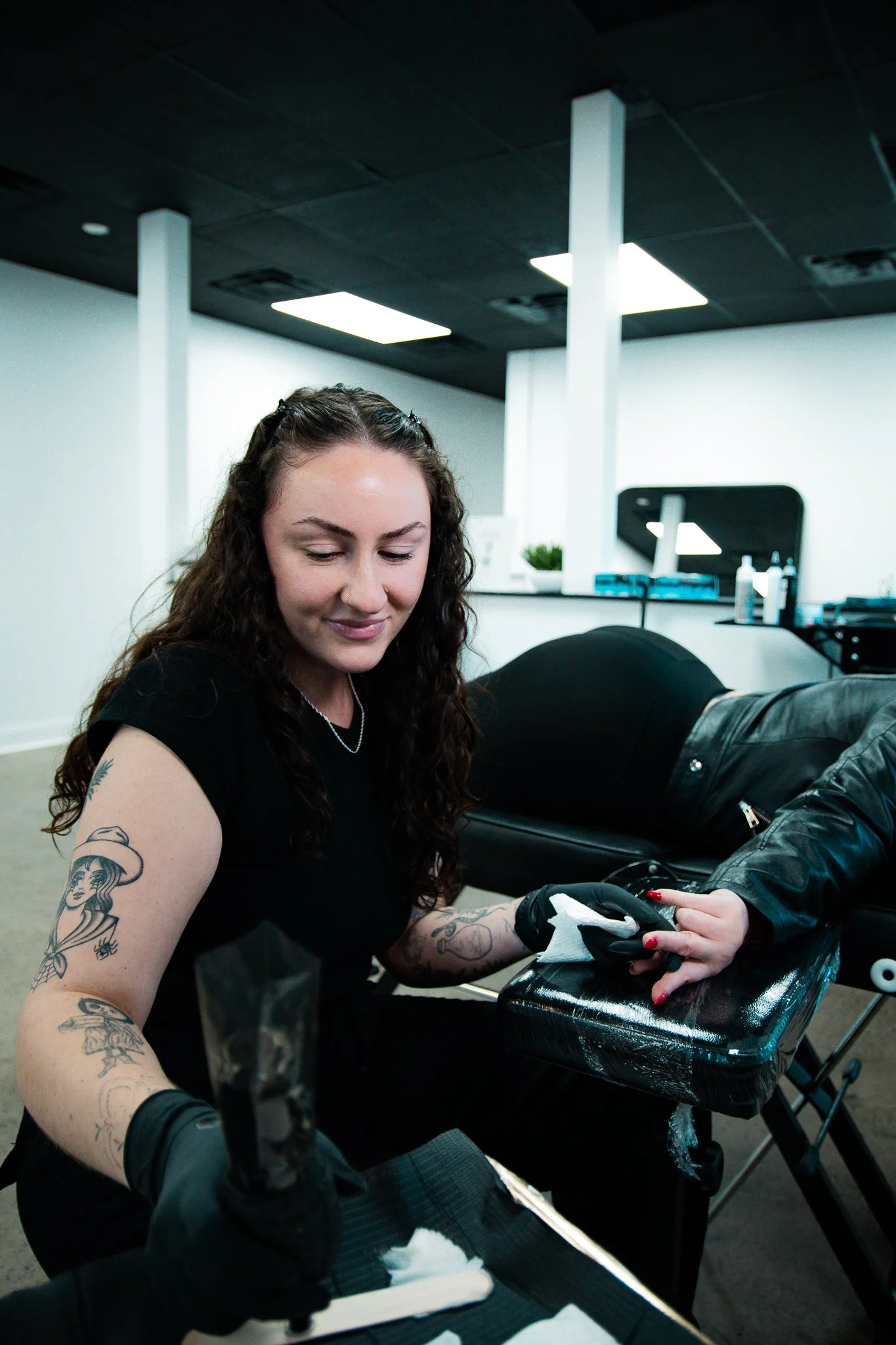 Tattoo artist with long curly hair and tattoos on her arms, wearing gloves, working on a client's arm in a tattoo studio.
