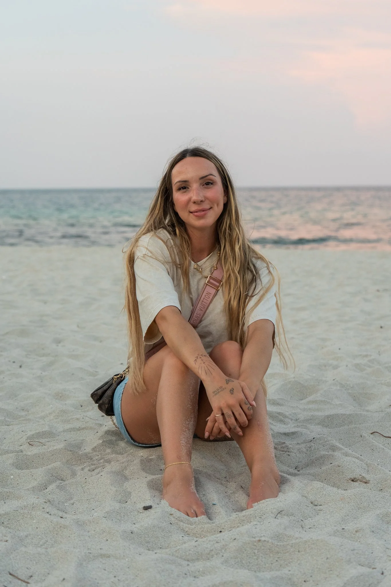 A young woman with long blonde hair sitting on a sandy beach at sunset, smiling at the camera, with the ocean and pink sky in the background.
