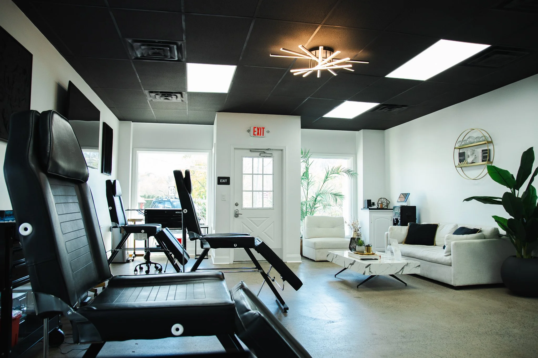 Interior of a modern waiting room with black gym chairs lined up, white sofas, a marble coffee table, potted plants, and contemporary lighting fixtures.