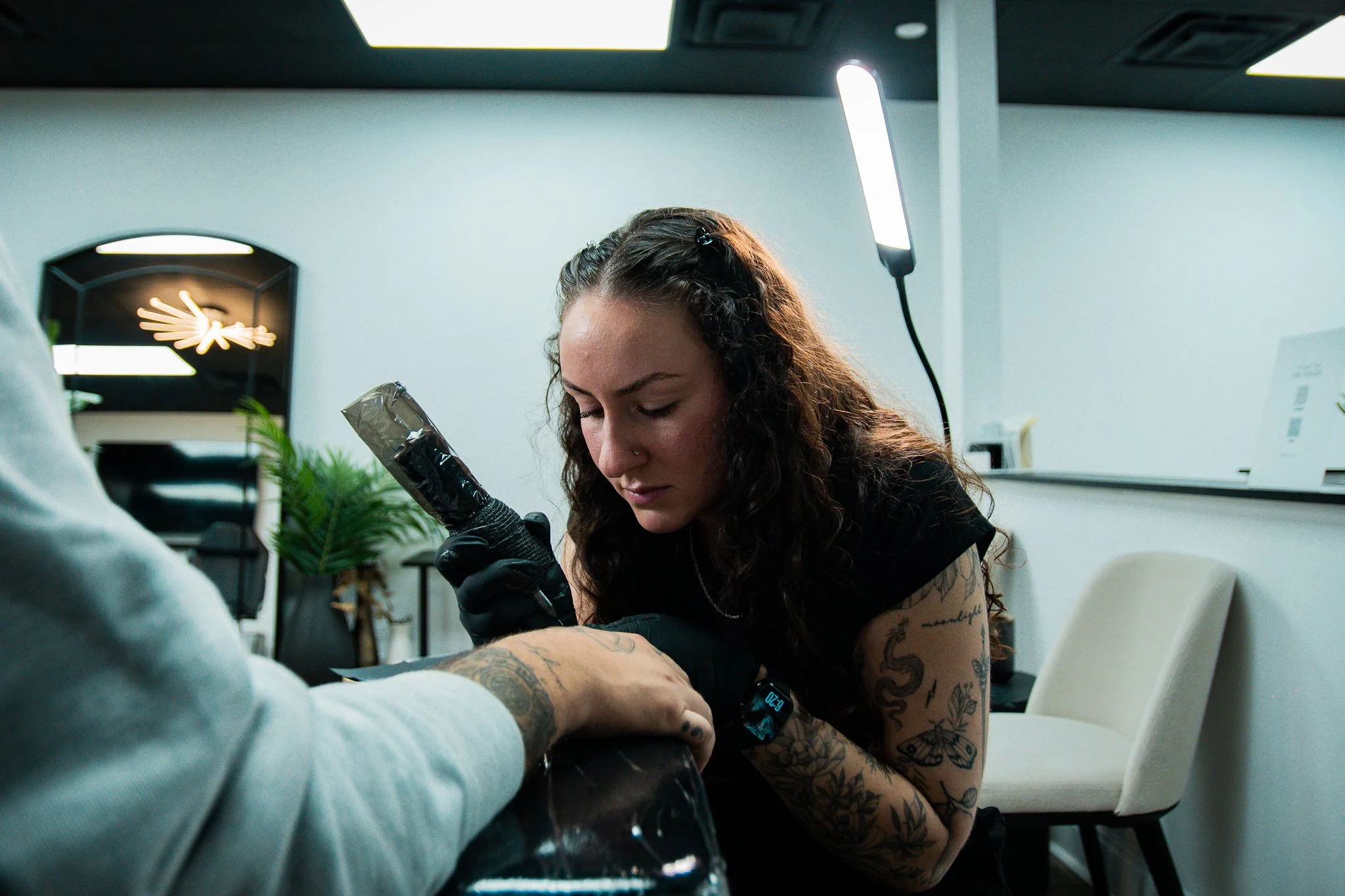 A woman with tattoos and curly hair getting a tattoo on her arm in a tattoo studio, with a tattoo artist working on her.