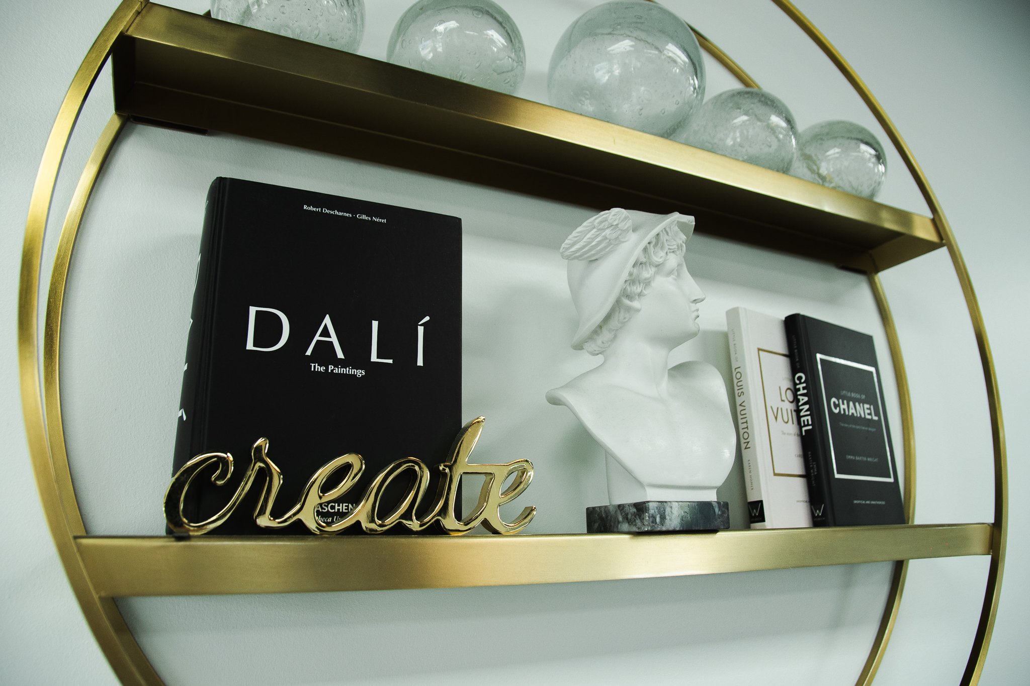 A gold-framed wall shelf holding decorative glass orbs, art books including 'Dalí: The Paintings' and Chanel, a white bust sculpture with a hat, and a gold 'create' sign