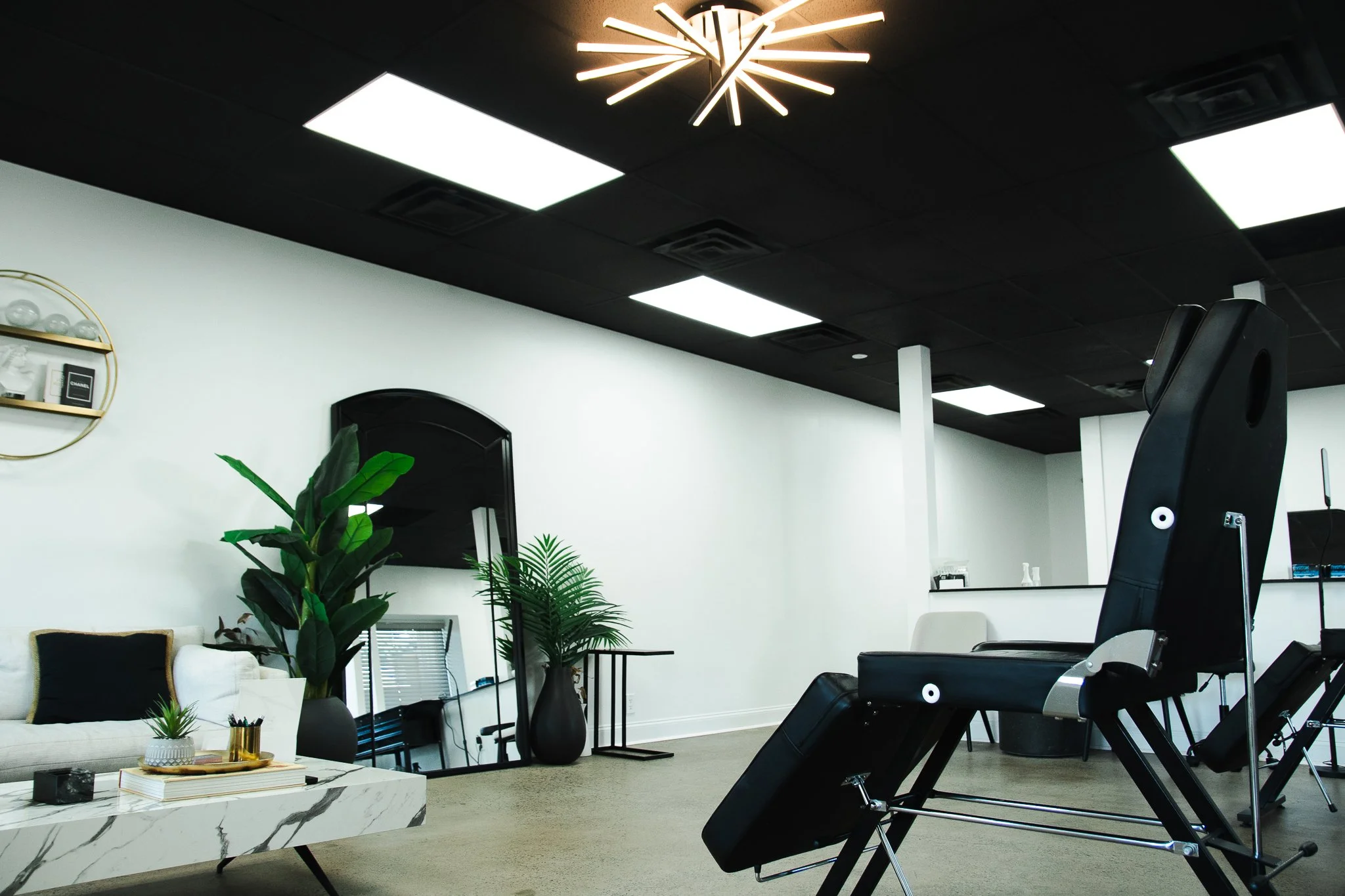A modern medical or spa treatment room with massage chair, sofa, plants, mirror, and decorative shelves under black ceiling with grid lights and a contemporary chandelier.