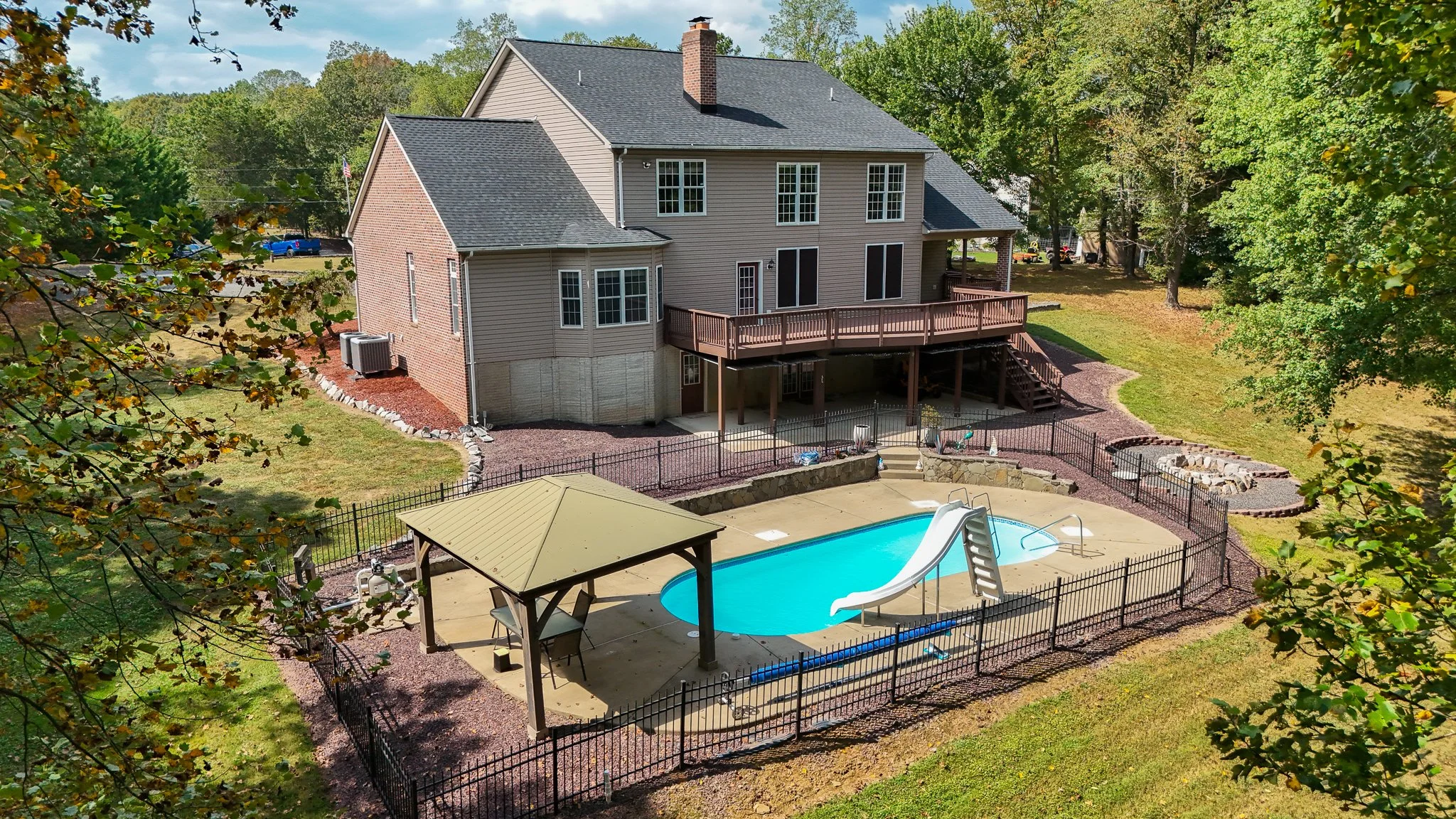 Backyard view of a two-story house with a swimming pool, slide, and pool house, surrounded by green trees.