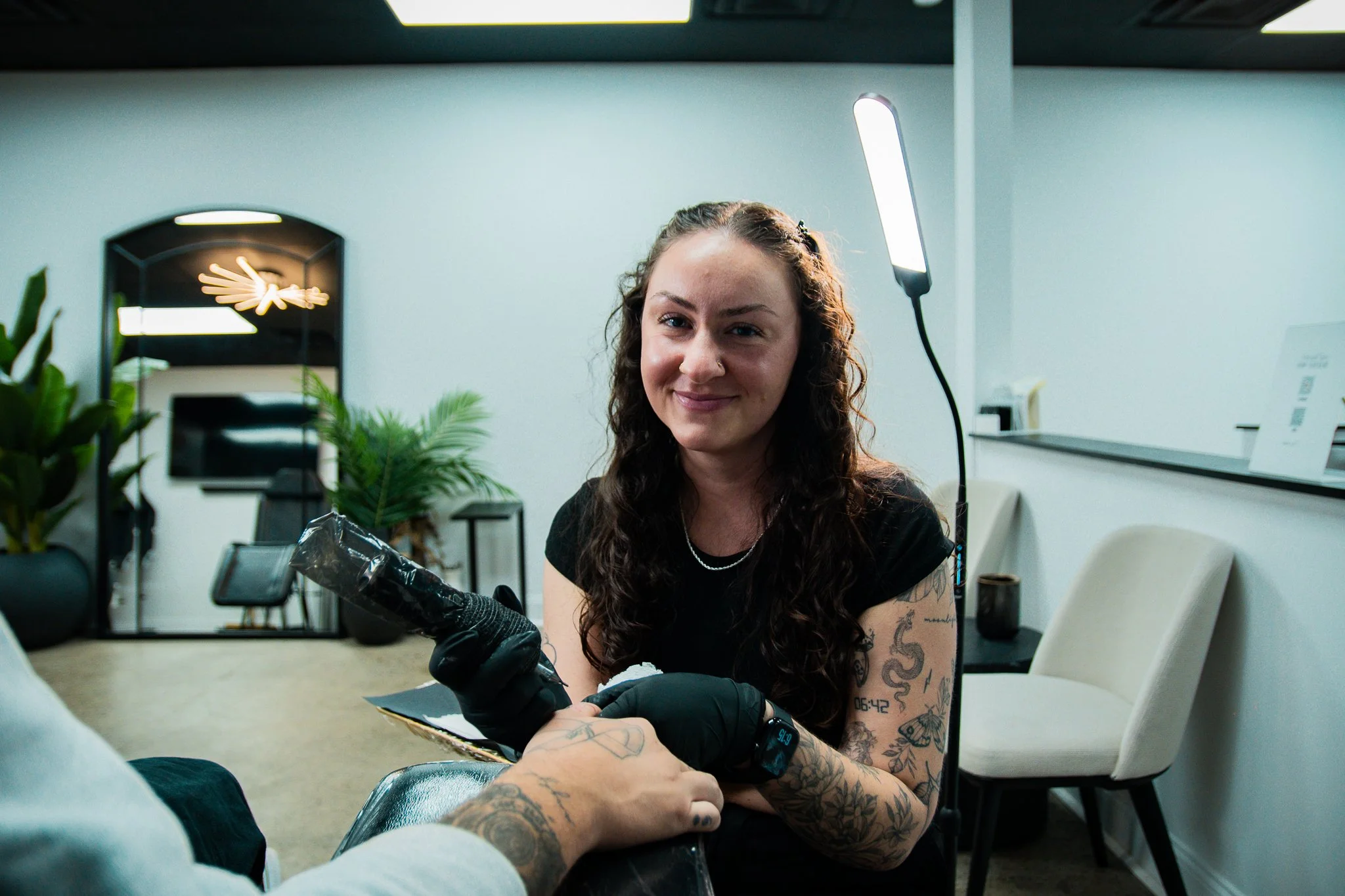 Tattooed woman sitting at a nail salon receiving a tattoo on her arm, smiling at the camera.