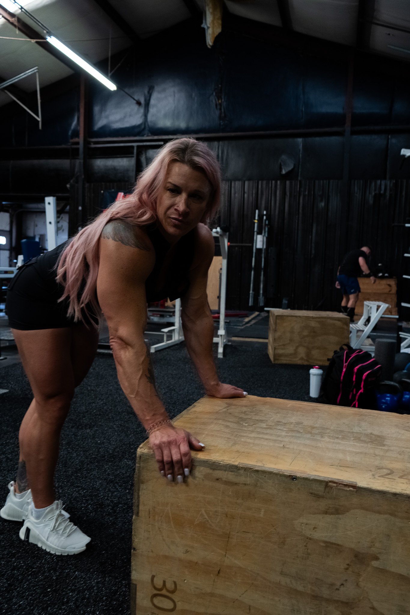 A woman with pink hair and tattoos performing a push-up on a wooden plyometric box inside a gym.