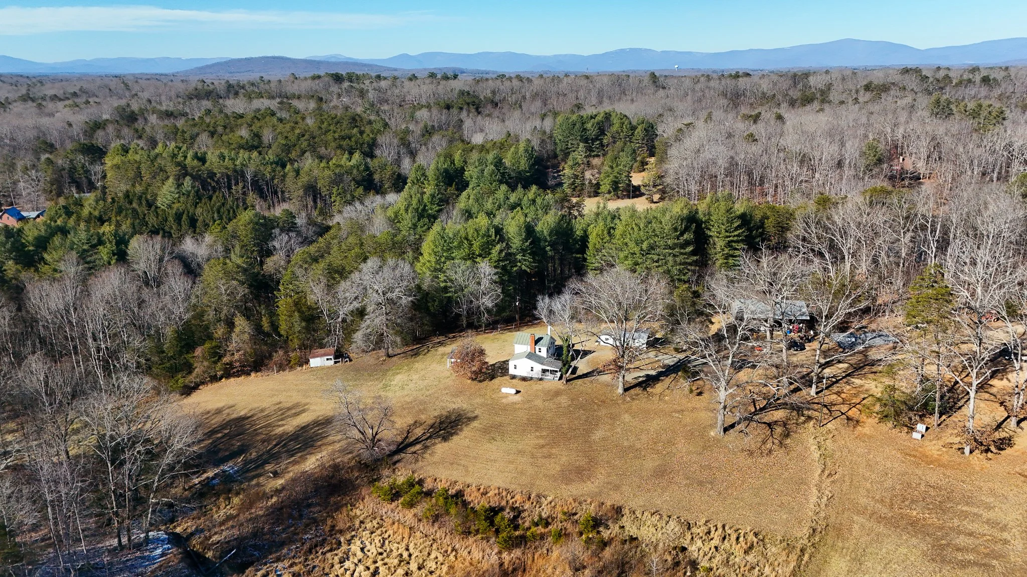 Aerial view of a rural property surrounded by woods and open land, showing a white house with a green roof, smaller outbuildings, and leafless trees, with distant mountains in the background.