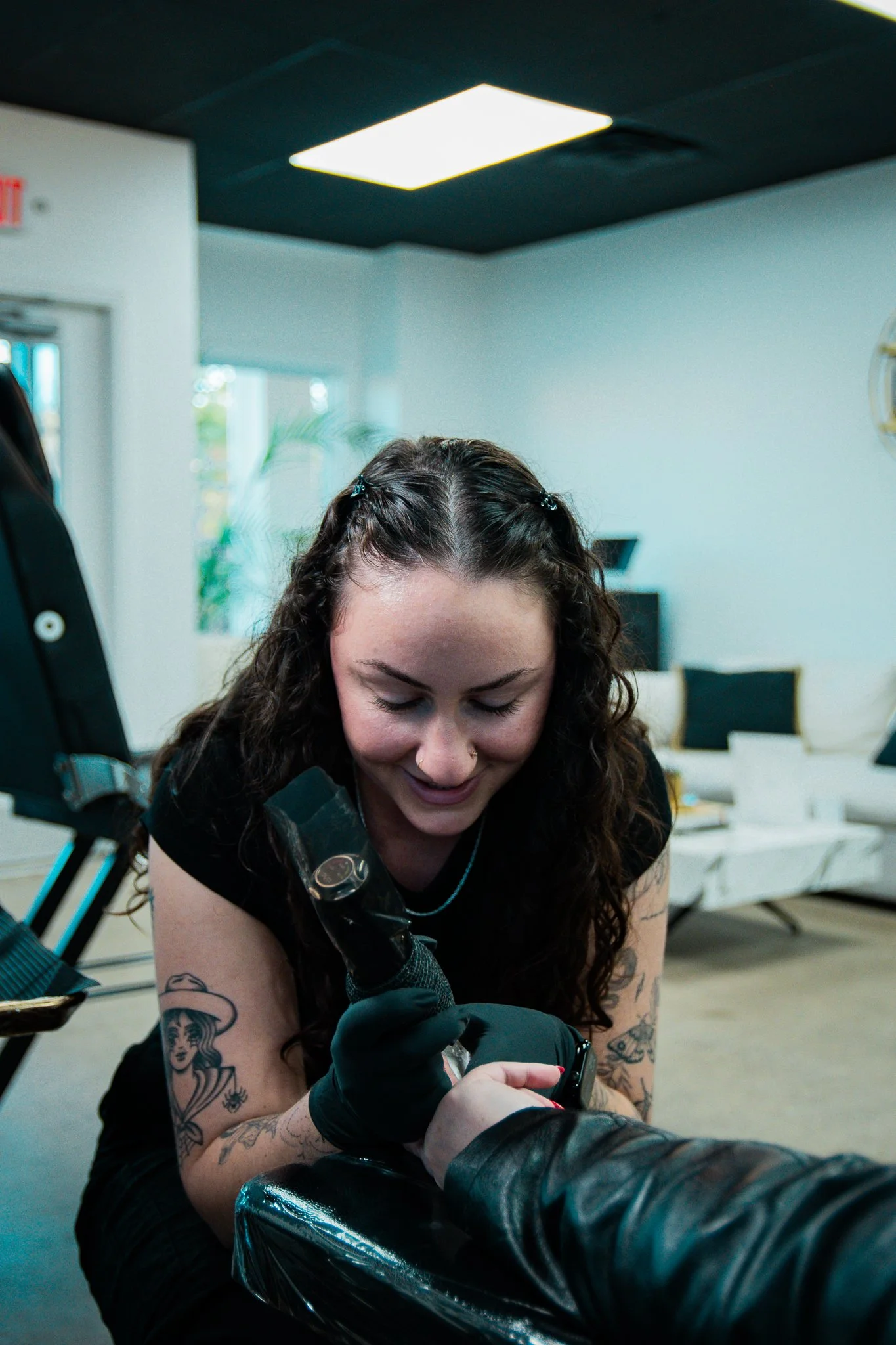 Tattooed woman getting a tattoo on her left hand in tattoo studio, with a black glove on her right hand, smiling and looking down.