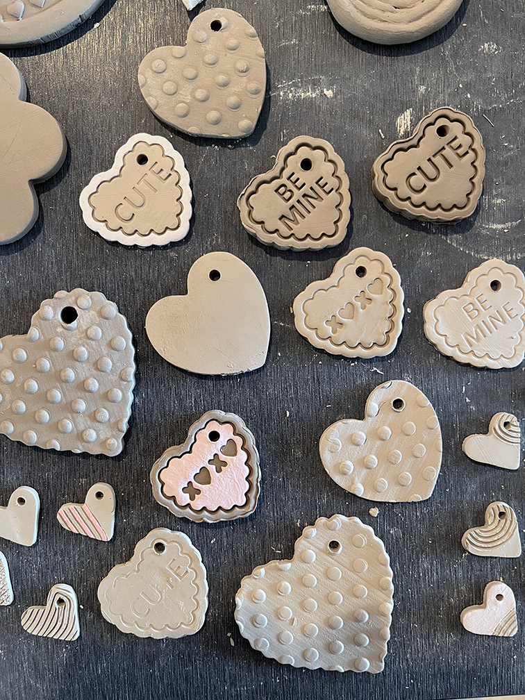 Various heart-shaped ceramic and rubber charms, some with textured patterns and inscriptions like "CUTE" and "BE MINE," laid out on a dark surface.