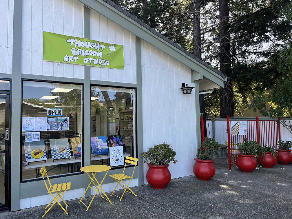 Exterior of an art studio called Thought Balloon Art Studio with a bright green sign, yellow outdoor furniture, and red planters, located in a tree-lined area.