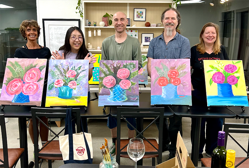Group of five people standing behind a table displaying five colorful paintings of flower bouquets in vases, with a lively and cheerful atmosphere.