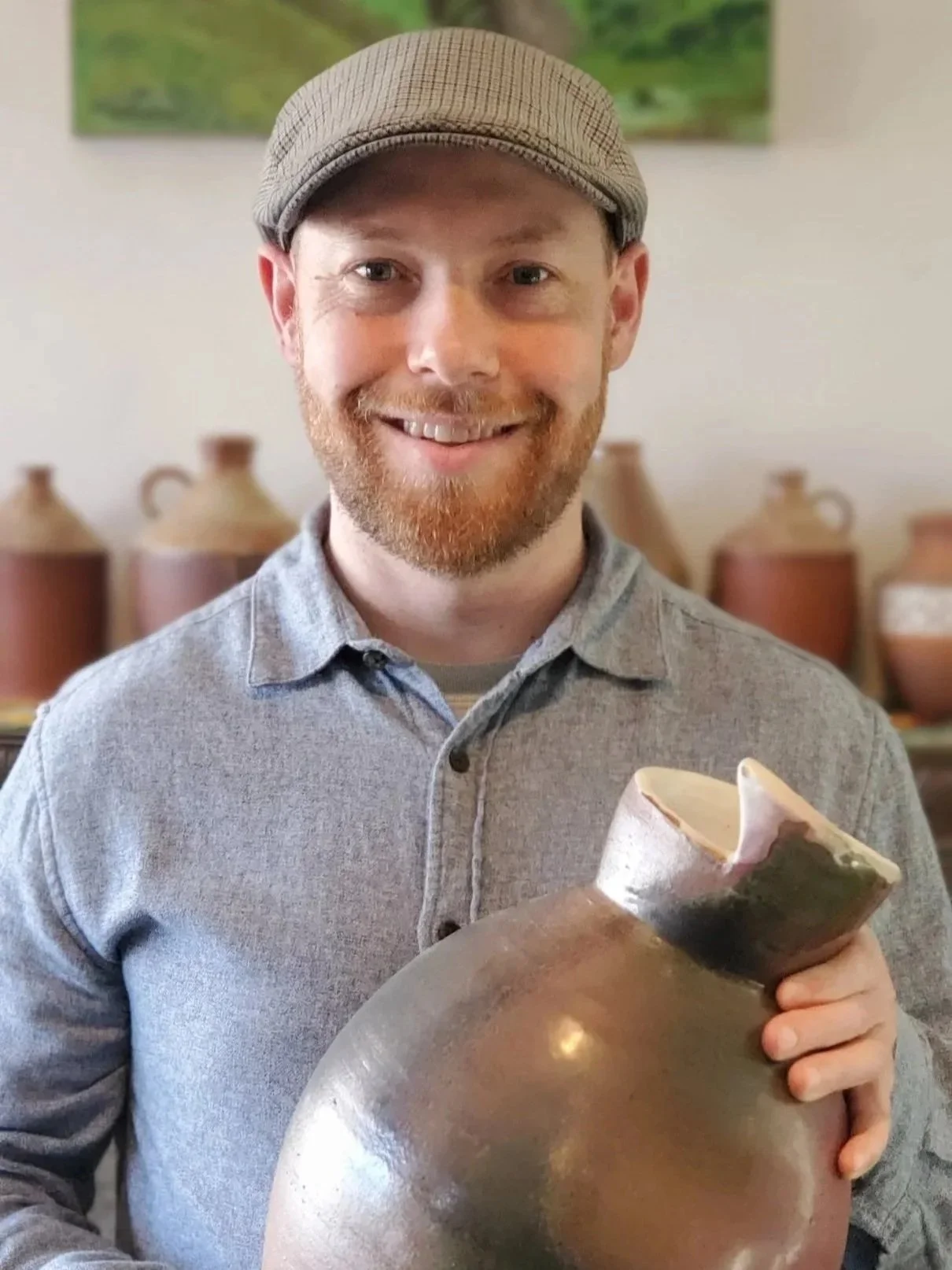 A smiling man wearing a gray shirt and a flat cap, holding a large ceramic vessel, with pottery jars in the background.
