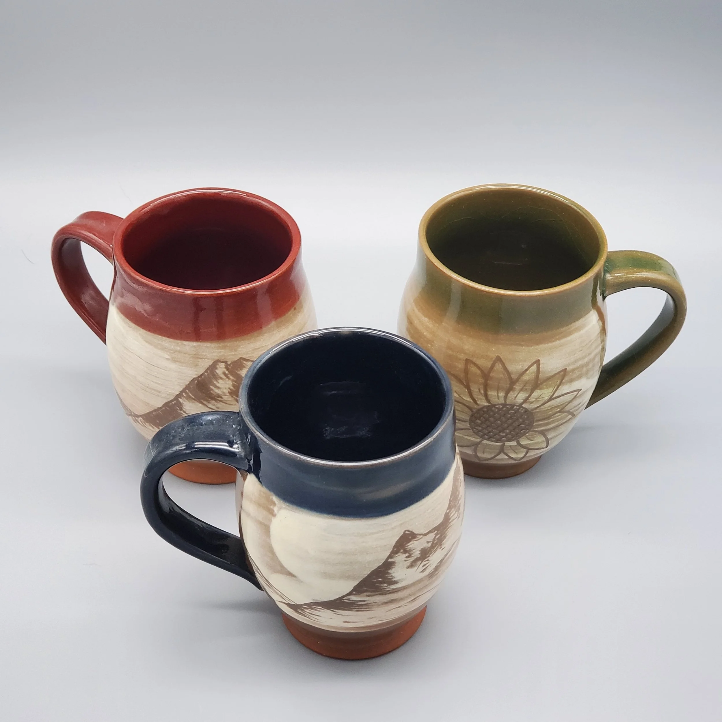 Three ceramic mugs with mountain and sunflower designs, each filled with a dark beverage, arranged on a white surface.