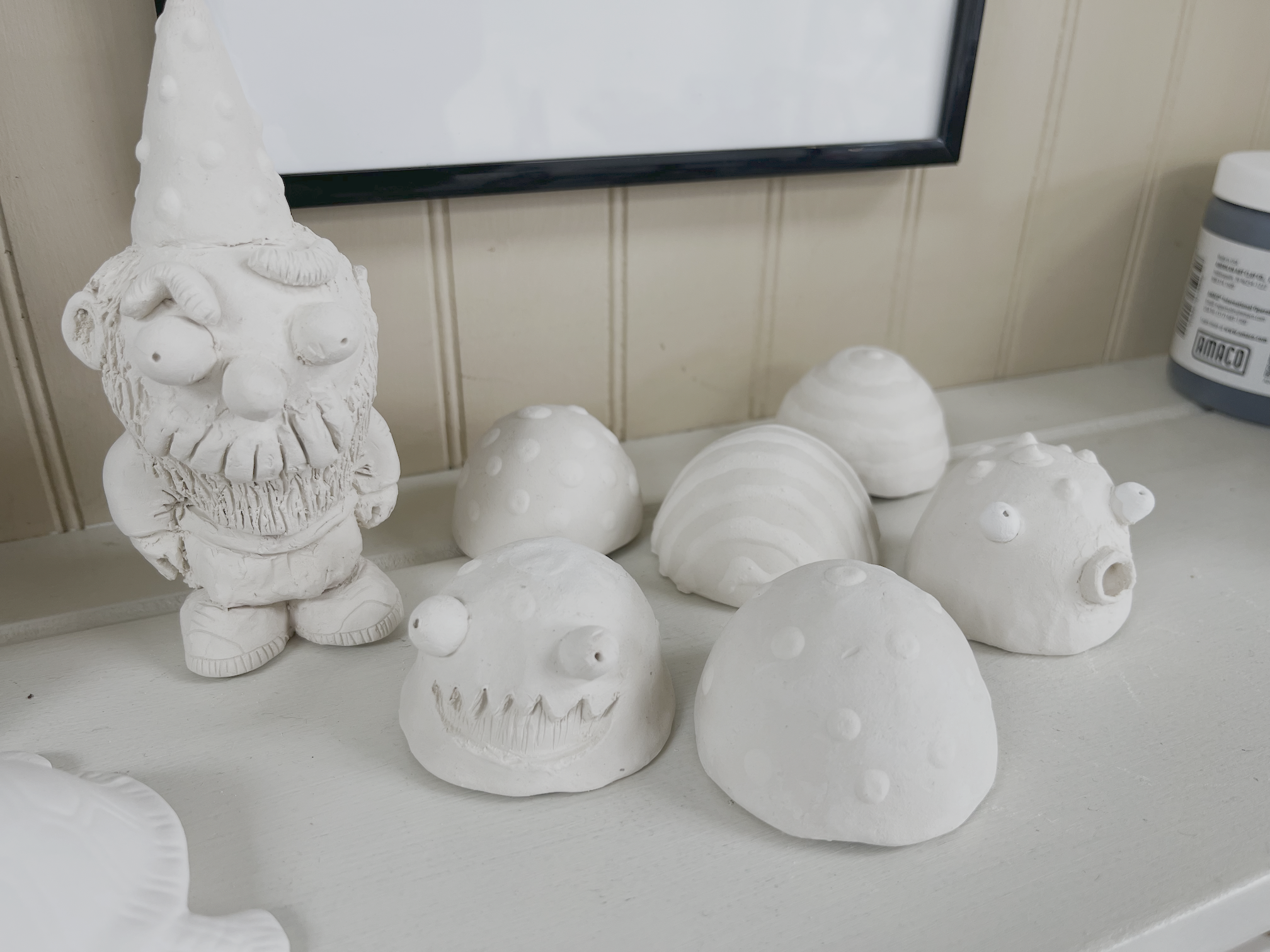 White ceramic gnome figurine with a pointed hat, beard, and striped shoes, surrounded by ceramic mushroom-shaped objects with various textured surfaces.
