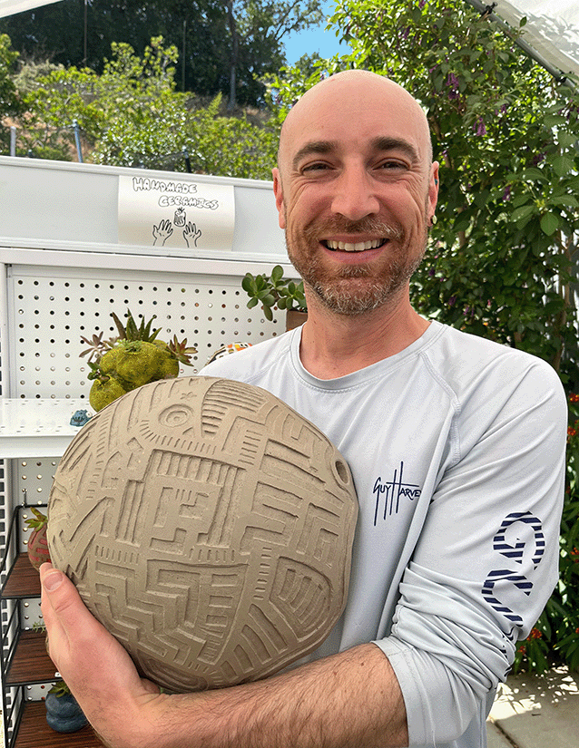 A man with a bald head and beard smiling, holding a large, spherical clay sculpture with intricate maze-like patterns. He is standing outdoors near a white shelf with plants and a sign that says "Handmade Ceramics."