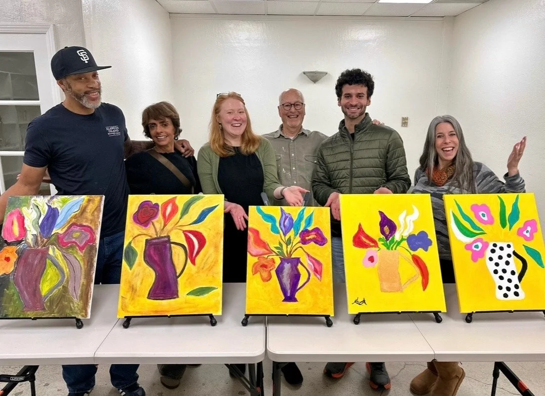 People standing behind tables displaying colorful abstract paintings of vases with flowers.