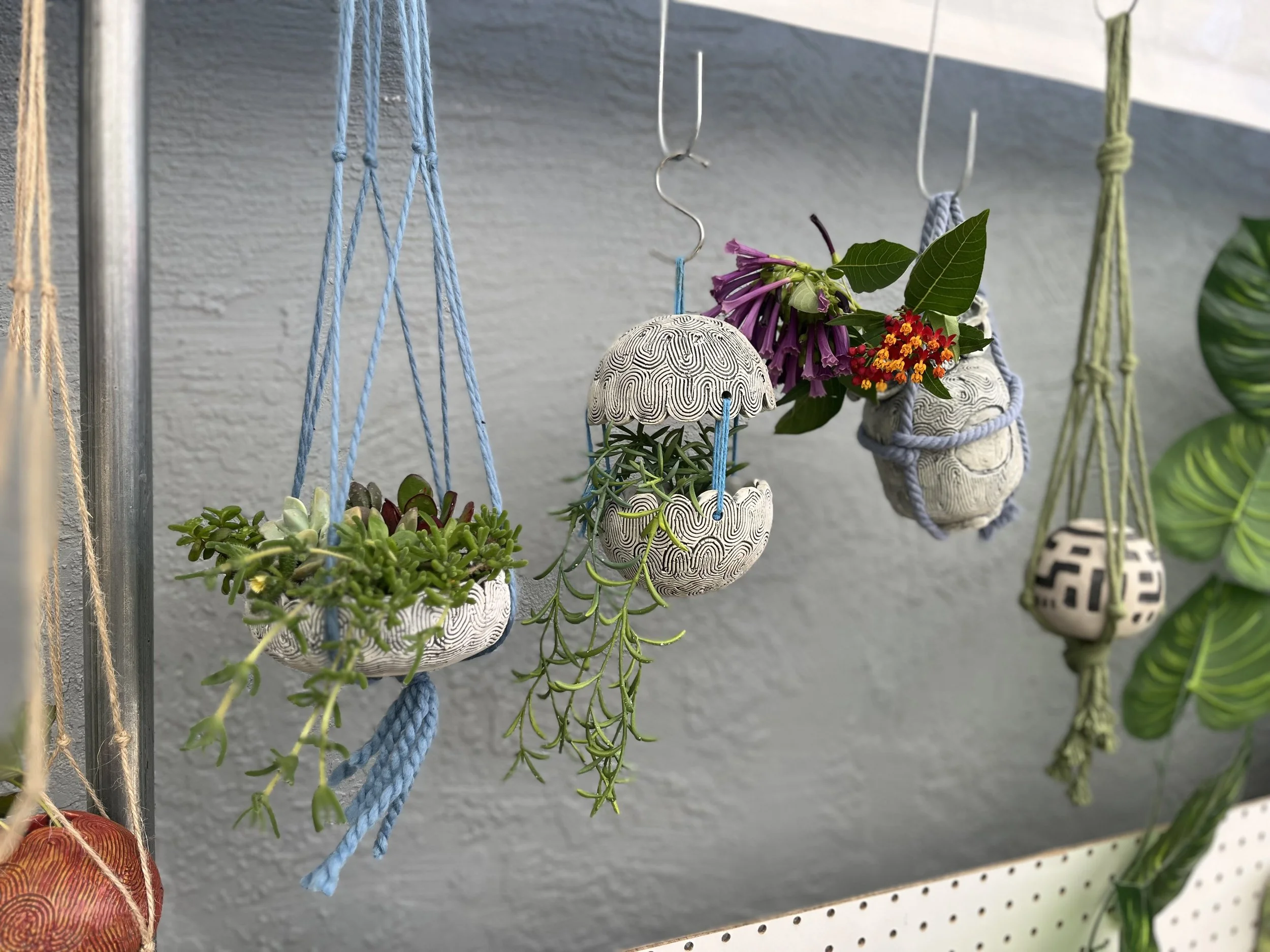 Decorative hanging planters with succulents and flowers, featuring artistic designs and colorful cords, displayed against a gray wall.