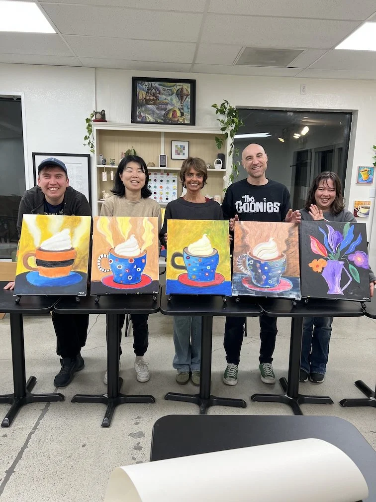 People standing behind tables with five colorful paintings of steaming cups of coffee or tea and a vase of flowers, in an indoor room.