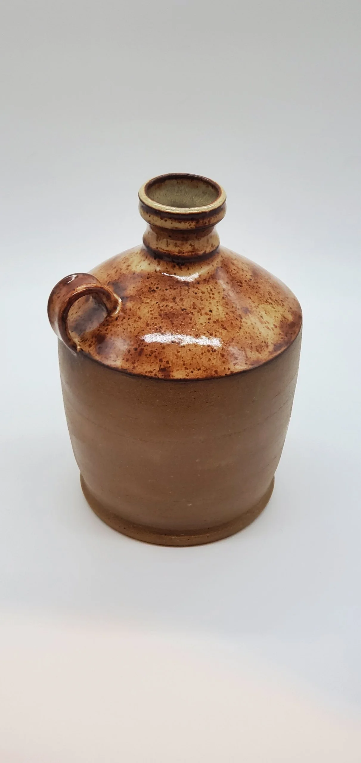 A ceramic pottery vessel with a round body, narrow neck, and small handle on the side, featuring a speckled brown glaze on the upper part and a matte, unglazed lower part.