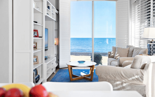 Hamptons style living room with grey linen sofas, a white and wooden coffee table, white wooden bookshelf and large panoramic views over the coean