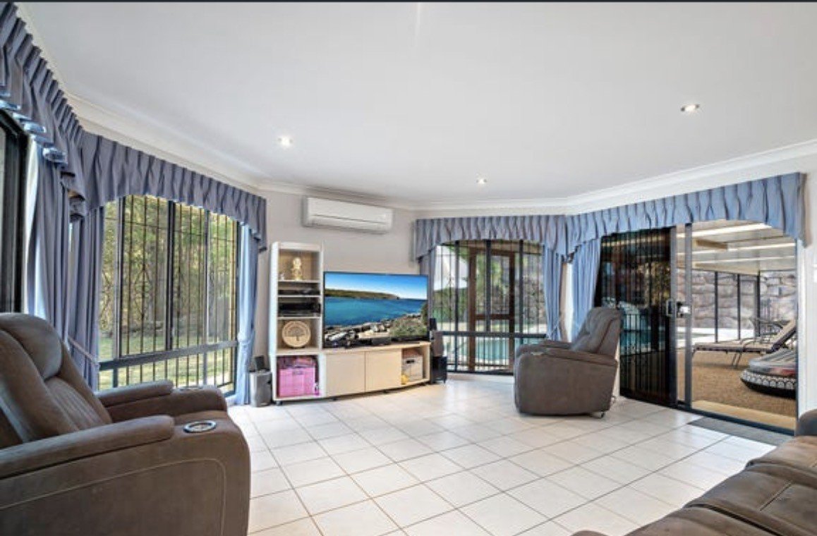 White tiled living room with dark brown leather couches, a TV and TV stand, and three large windows with purple curtains