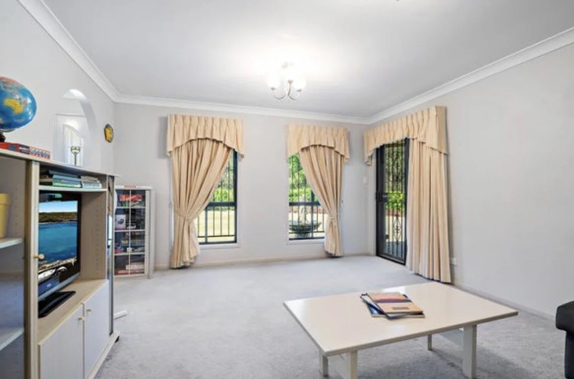 living room with grey carpet, a TV stand and TV off to the right, three large windows with beige curtains