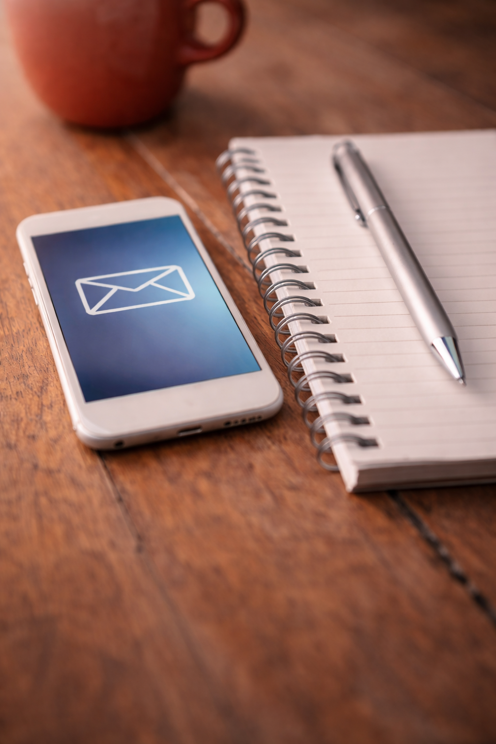 Smartphone with email icon, spiral notepad, silver pen, and red mug on wooden table.