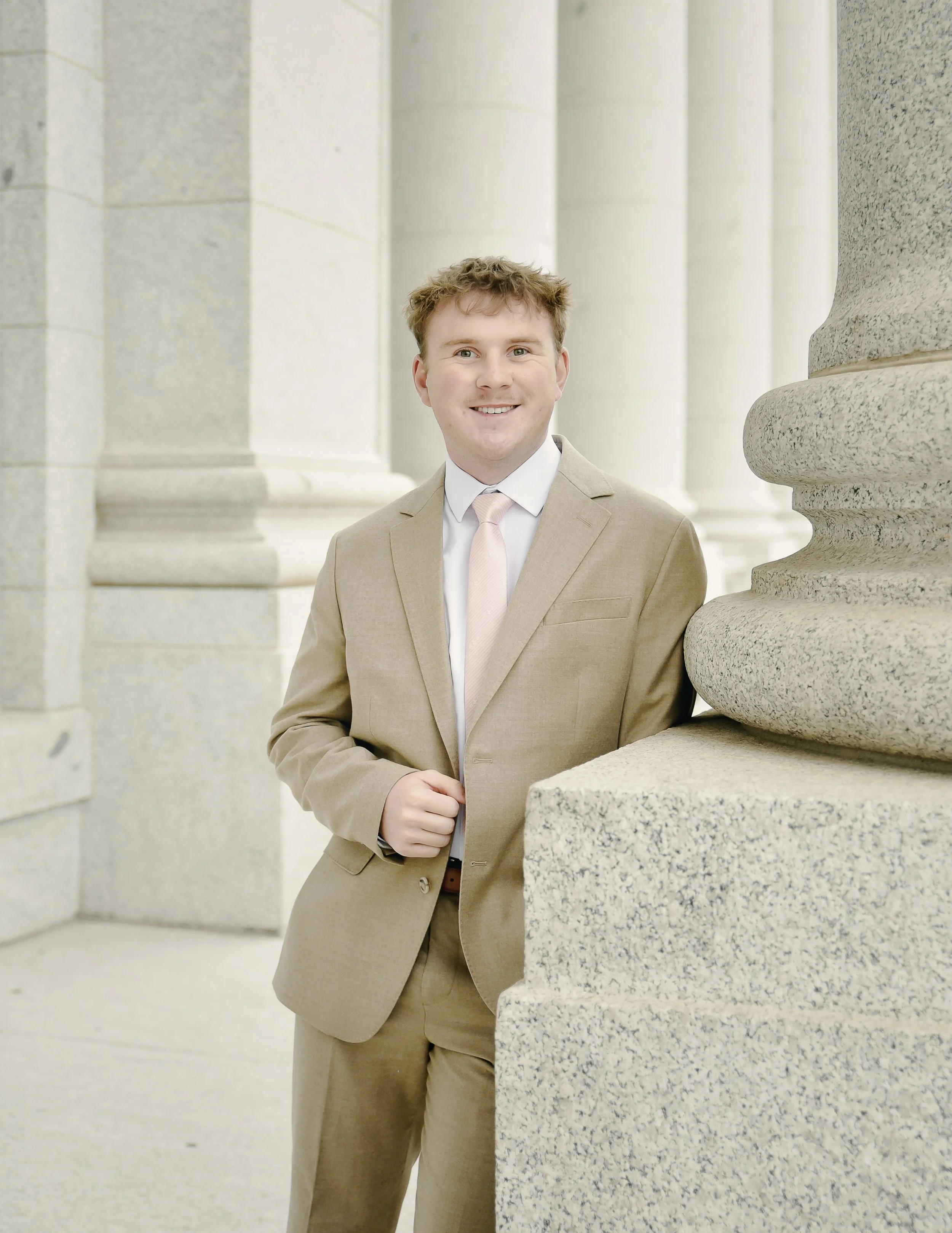 A young man in a beige suit and pink tie standing outside a stone building with large columns.