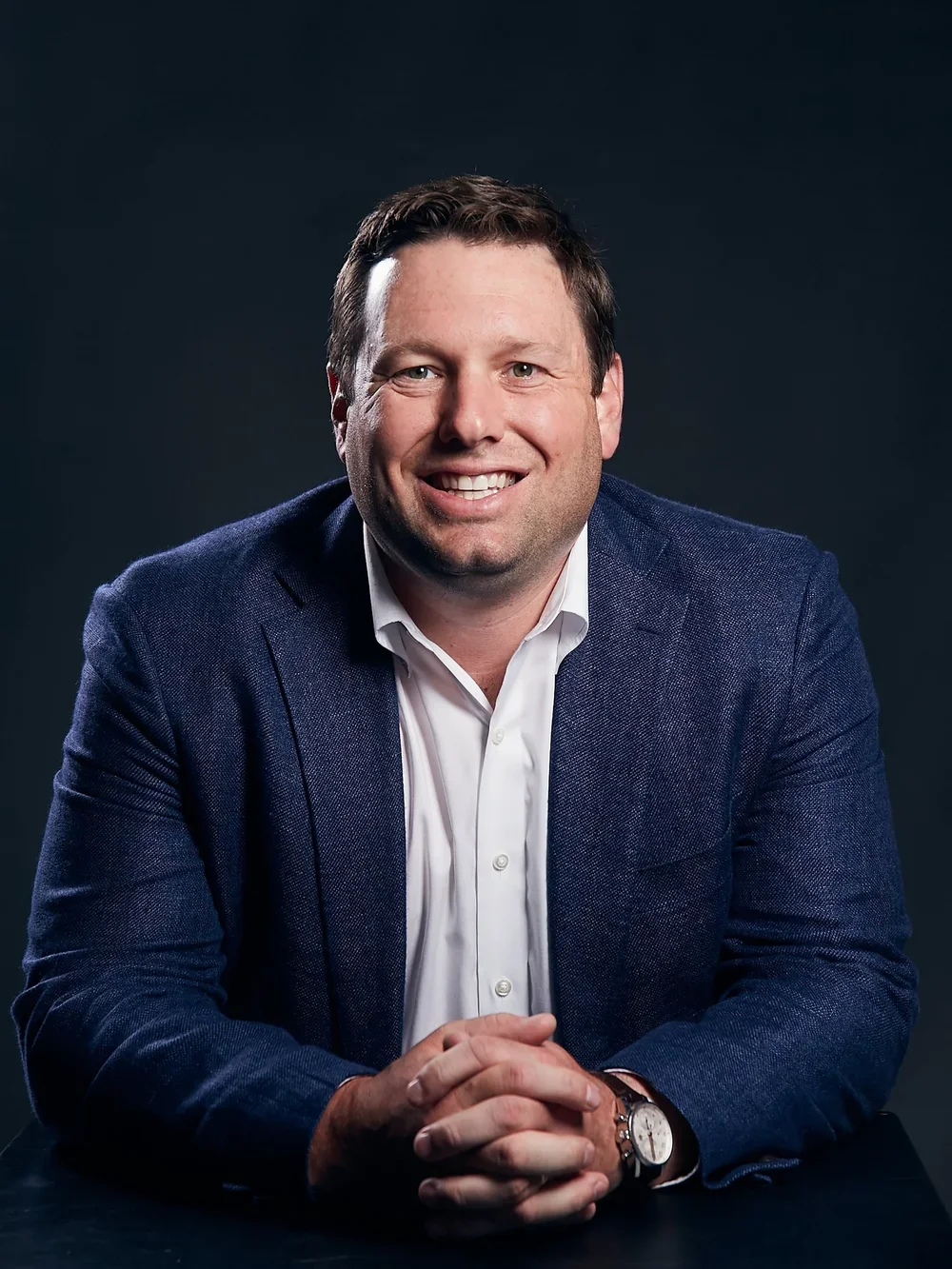 A man smiling, wearing a dark blue blazer, white shirt, and a wristwatch, sitting against a dark background.