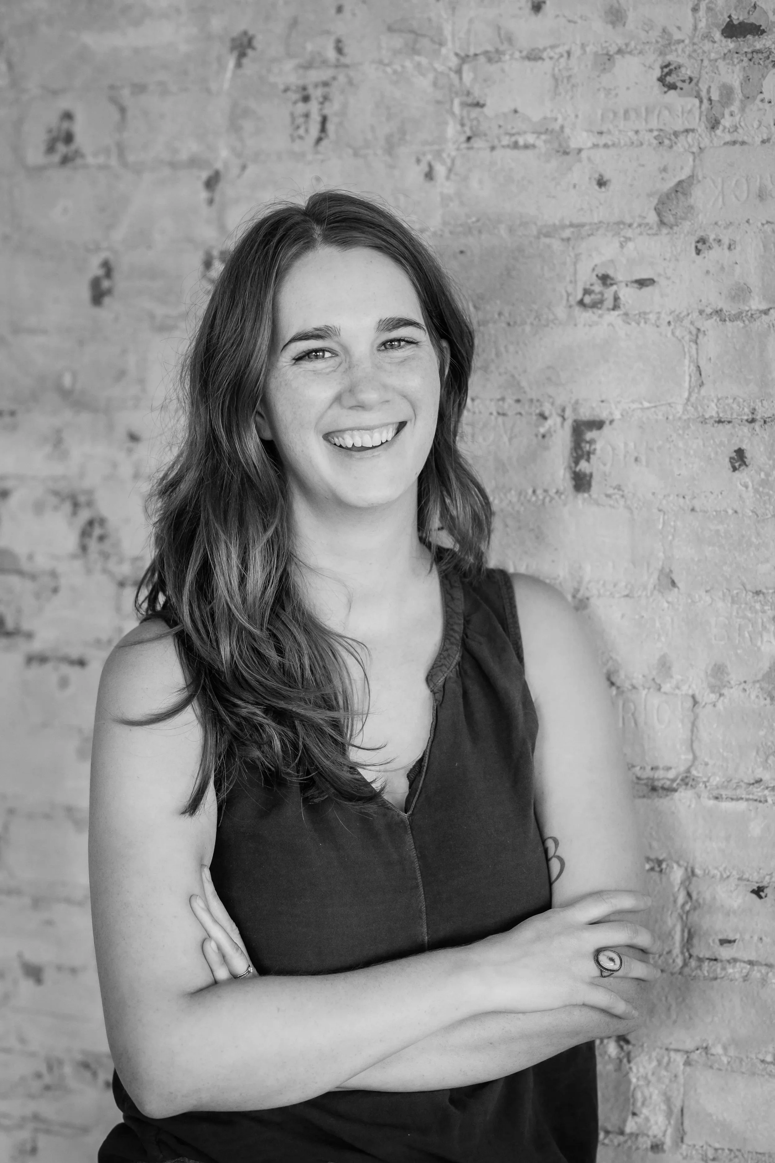 Black and white portrait of a smiling woman with long wavy hair, crossed arms, wearing a sleeveless top, standing against a brick wall.