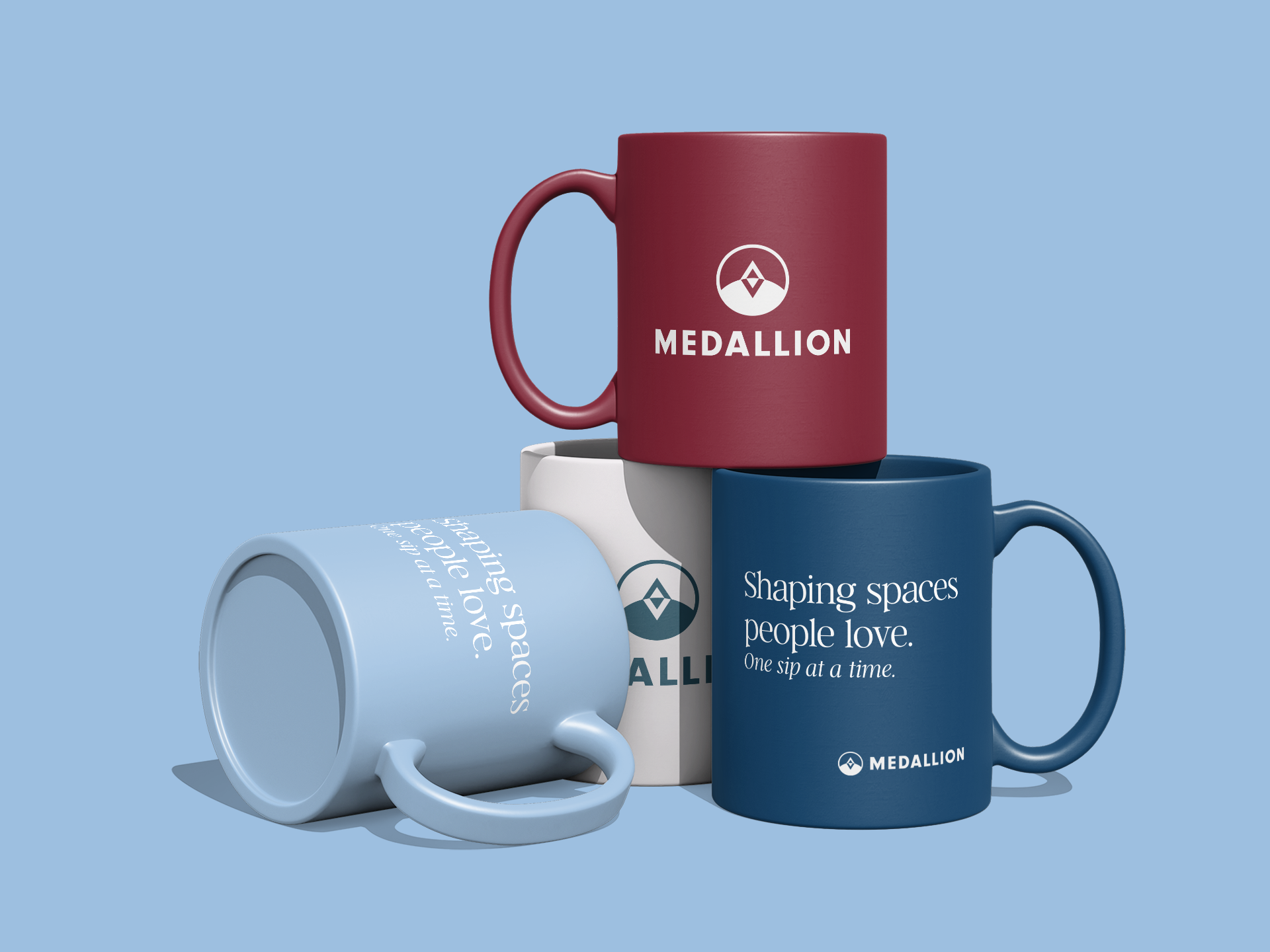 Three colored coffee mugs with Medallion branding, one red, one blue, and one white, arranged on a light blue background. The blue mug features the text "Shaping spaces people love. One sip at a time." The white mug is lying on its side, and the red 