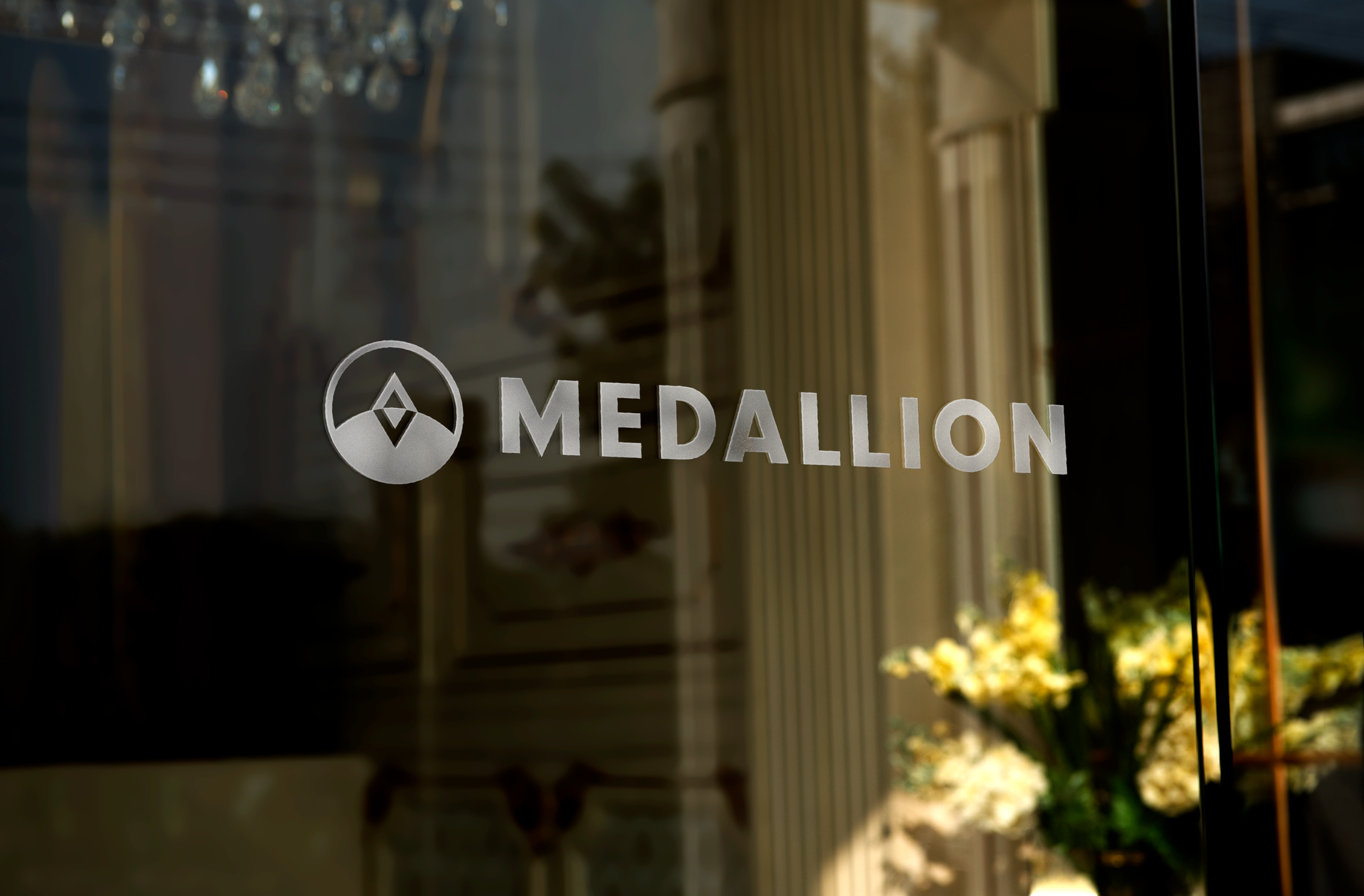 The glass door of a store or business with the logo and name 'MEDALLION' printed on it, reflecting the outdoor surroundings including flowers and a building.