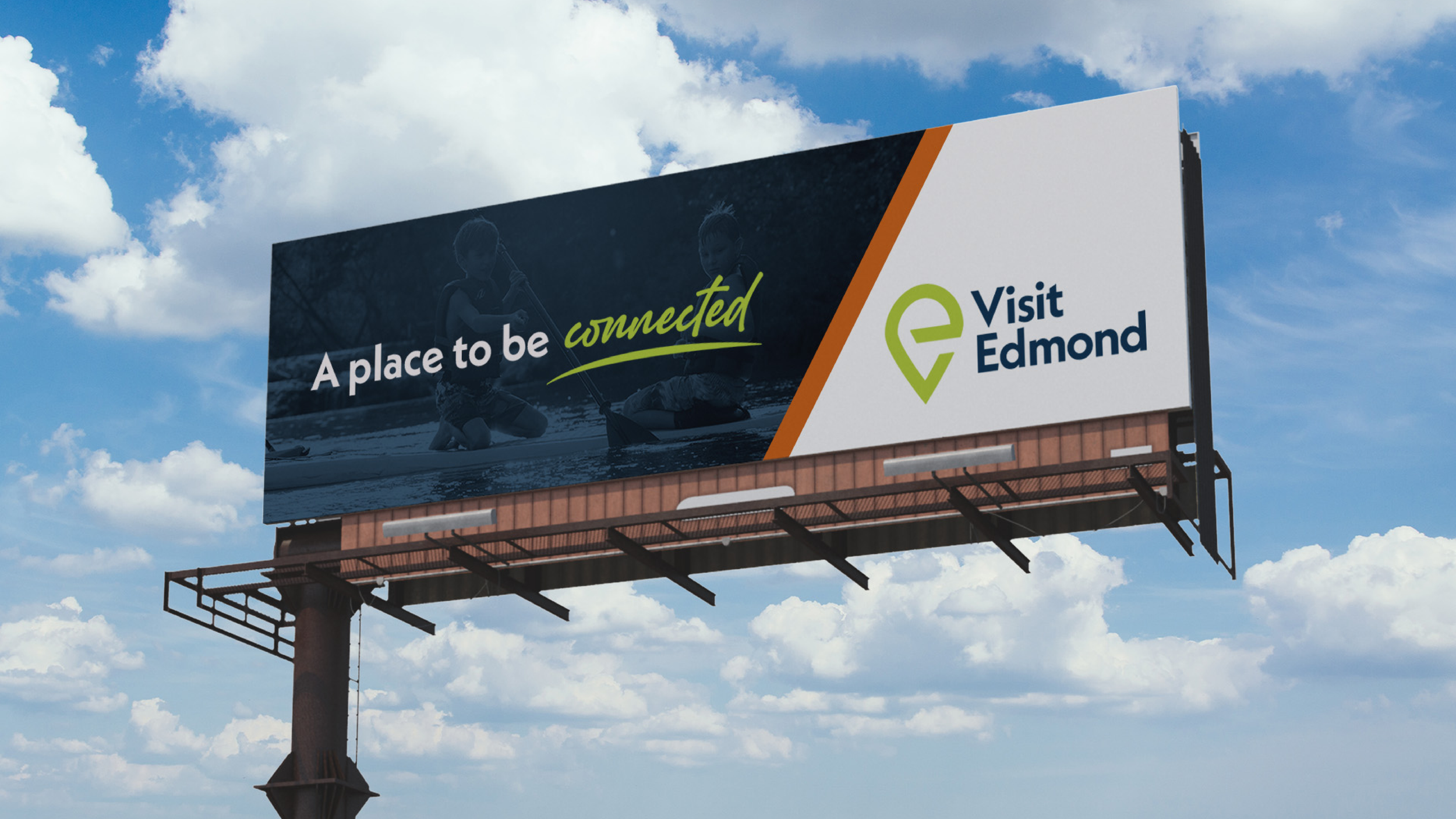 Billboard advertisement with a dark background on the left featuring two children playing by the water and the text "A place to be connected". The right side has a white background with the logo and text "Visit Edmond" in green and dark blue.