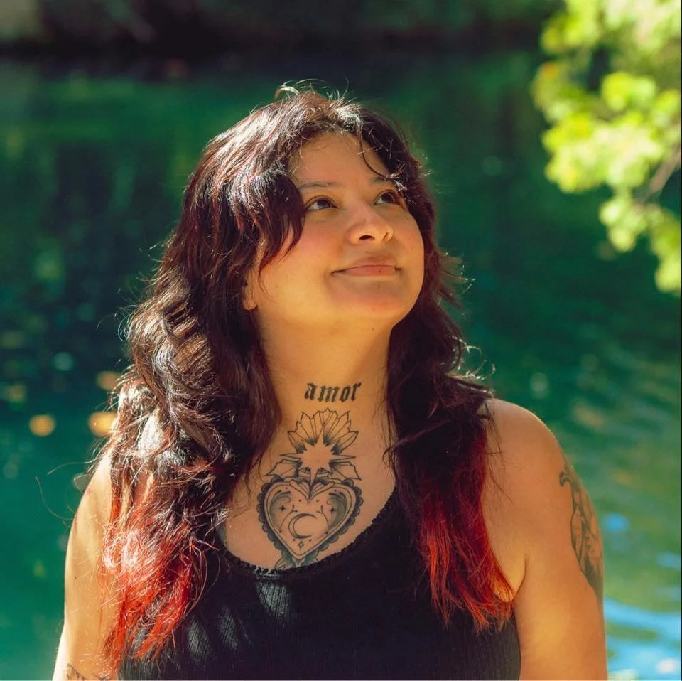 A woman with wavy dark hair and red tips smiling outdoors near a body of water with green foliage in the background.