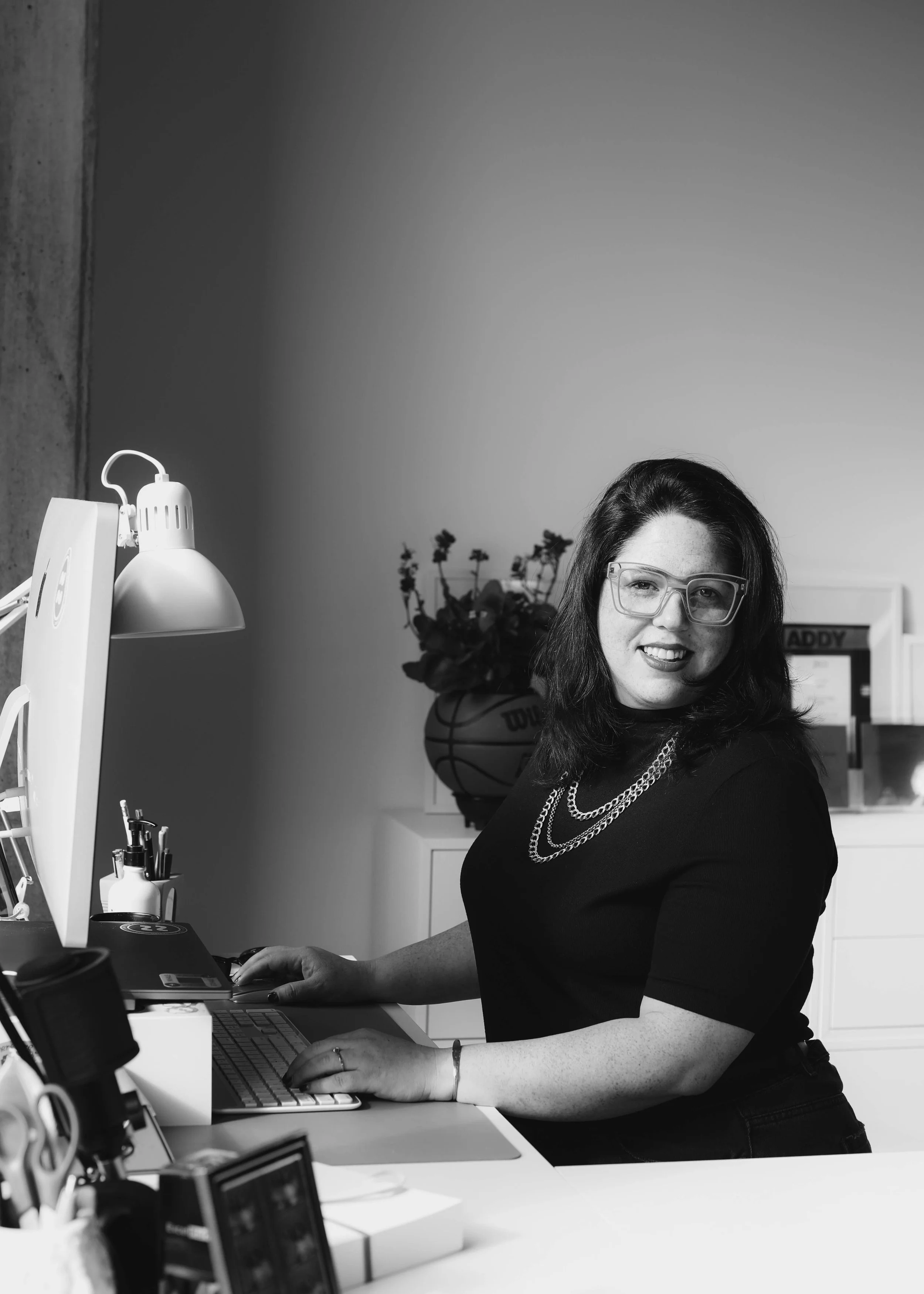 Black-and-white photo of a woman with shoulder-length dark hair, glasses, wearing a black top and necklaces, sitting at a desk with a computer. There are office supplies, a lamp, and a basketball with flowers in the background.