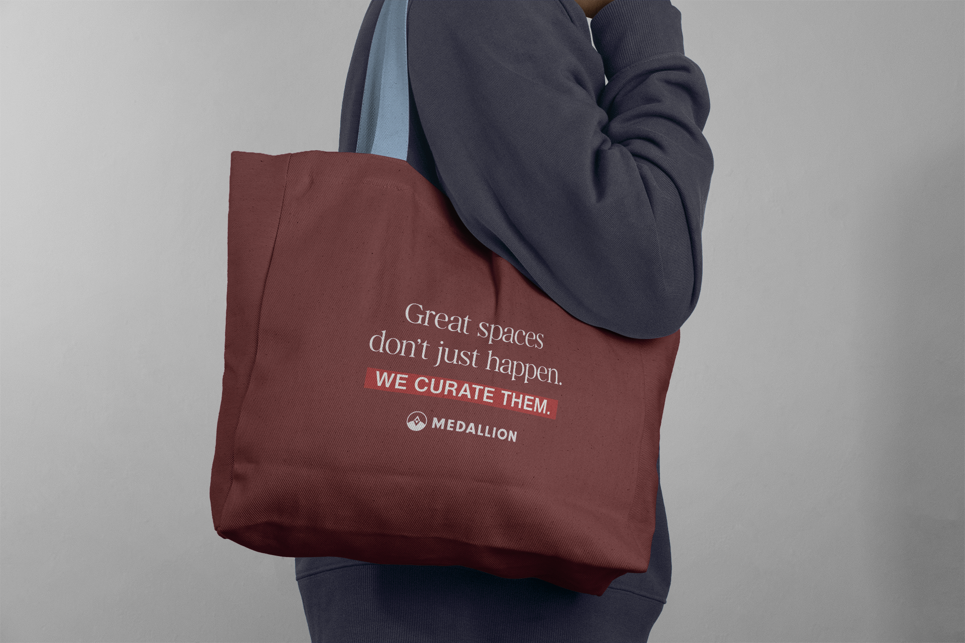 Person wearing a dark blue sweatshirt and carrying a large maroon tote bag with white and red text that says, "Great spaces don't just happen. WE CURATE THEM." and a logo underneath.