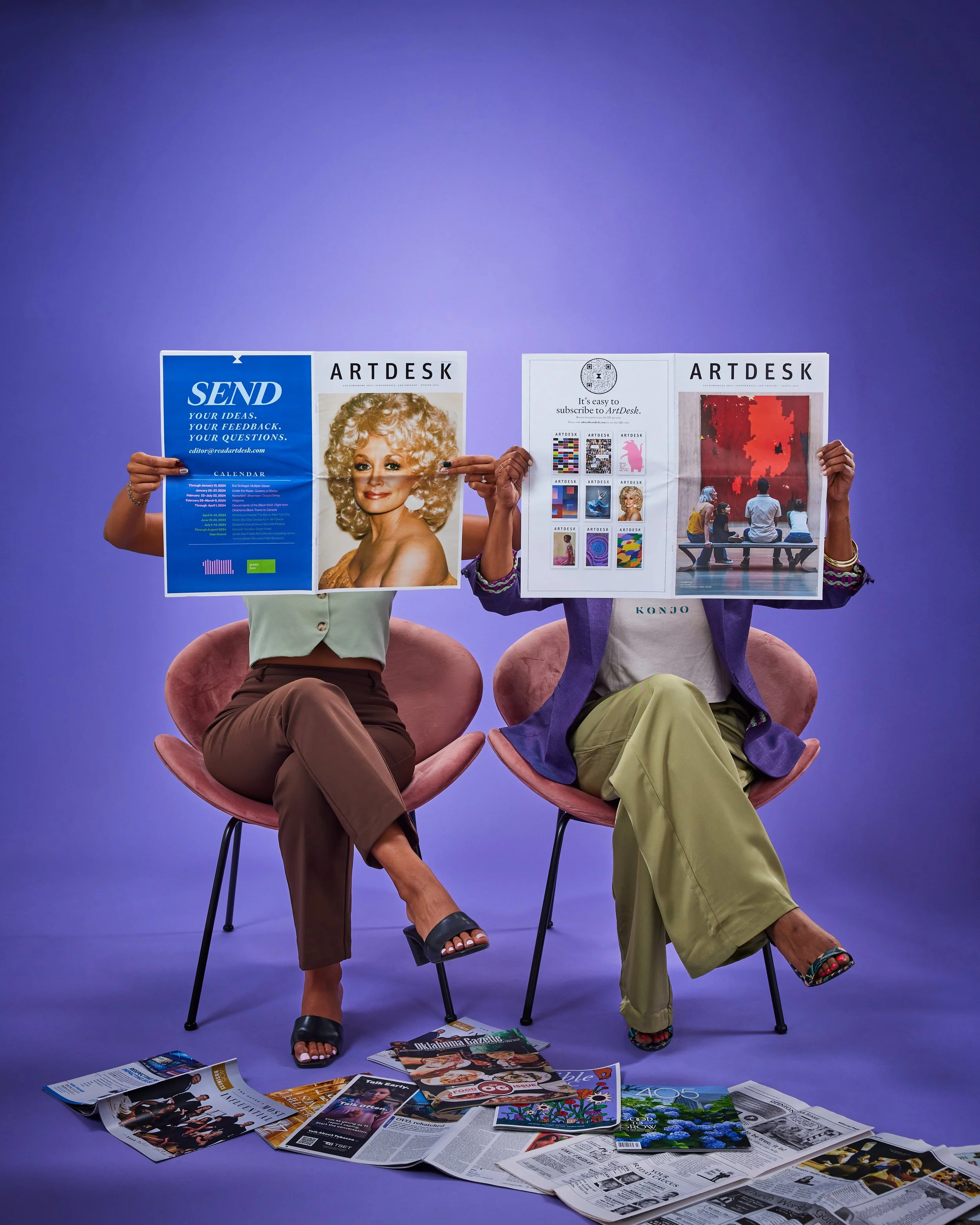 Two women sitting on pink chairs holding up magazines in front of their faces. The woman on the left wears a mint green top and brown pants, the woman on the right wears a purple jacket and khaki pants. Magazines and newspapers are scattered on the floor.