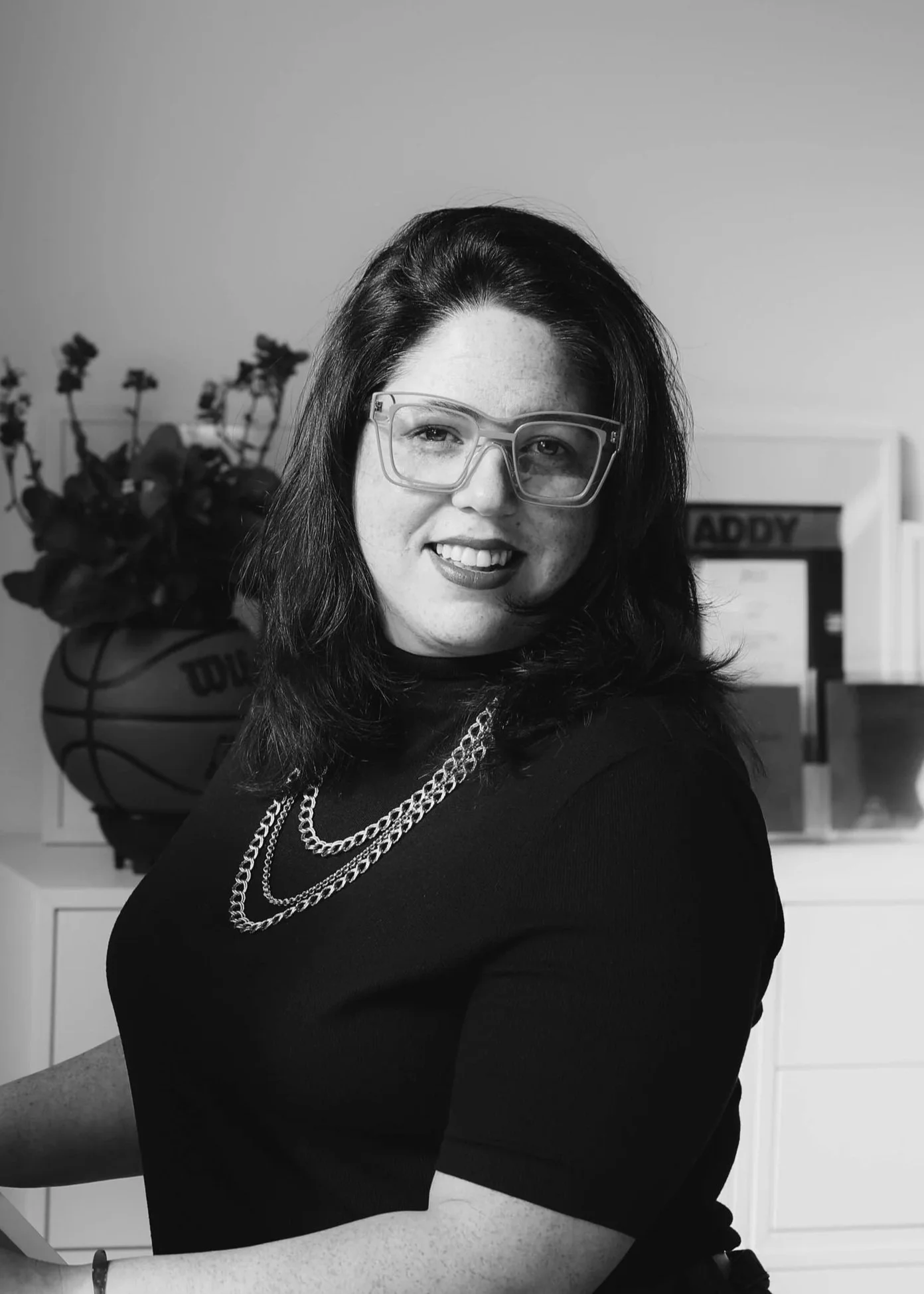 A woman with glasses and dark hair, smiling and wearing layered necklaces, sitting in an indoor setting with a basketball and potted plant in the background.