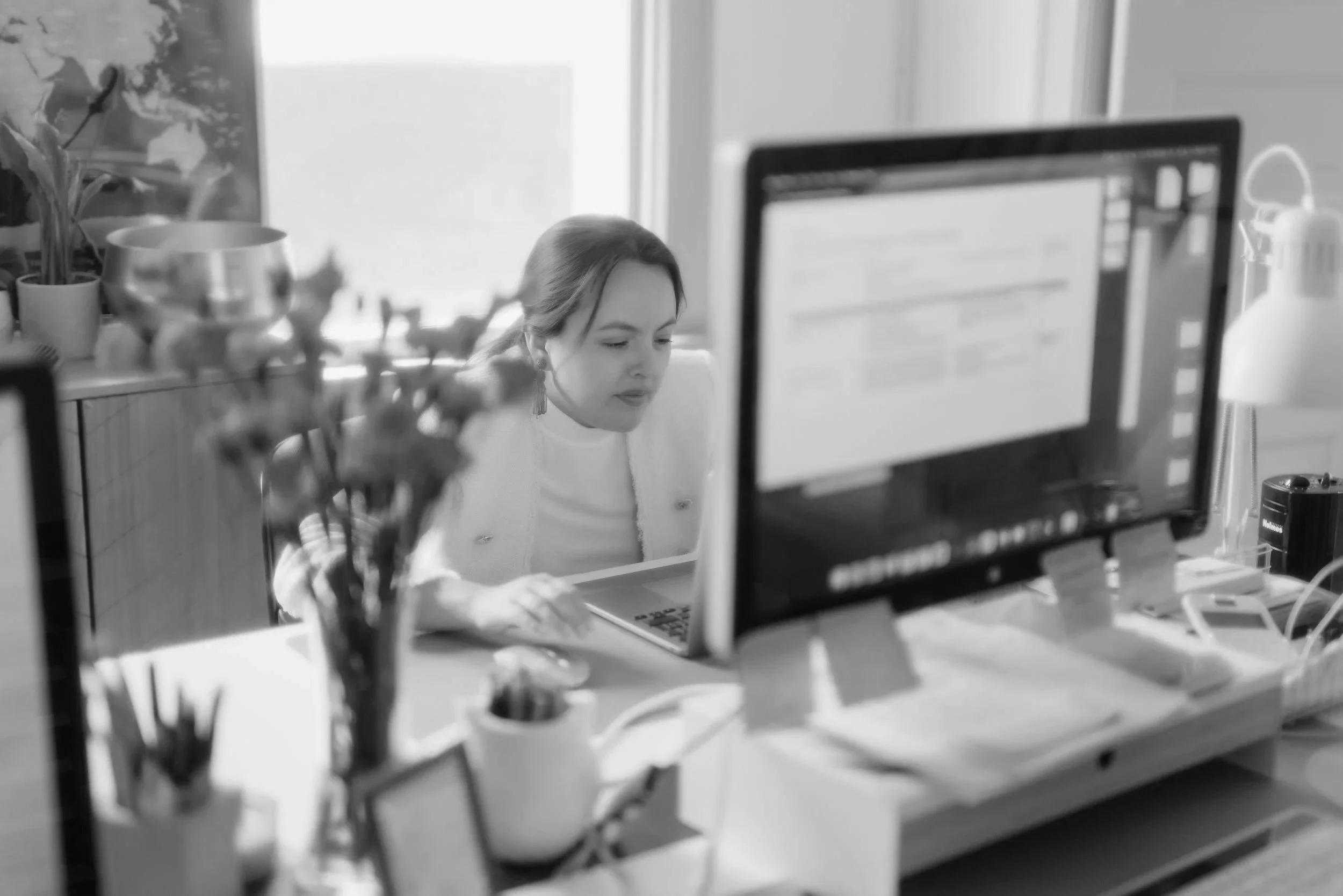 A woman sitting at a cluttered office desk, working on a laptop with a computer monitor in front of her, in a room with a window and plant.