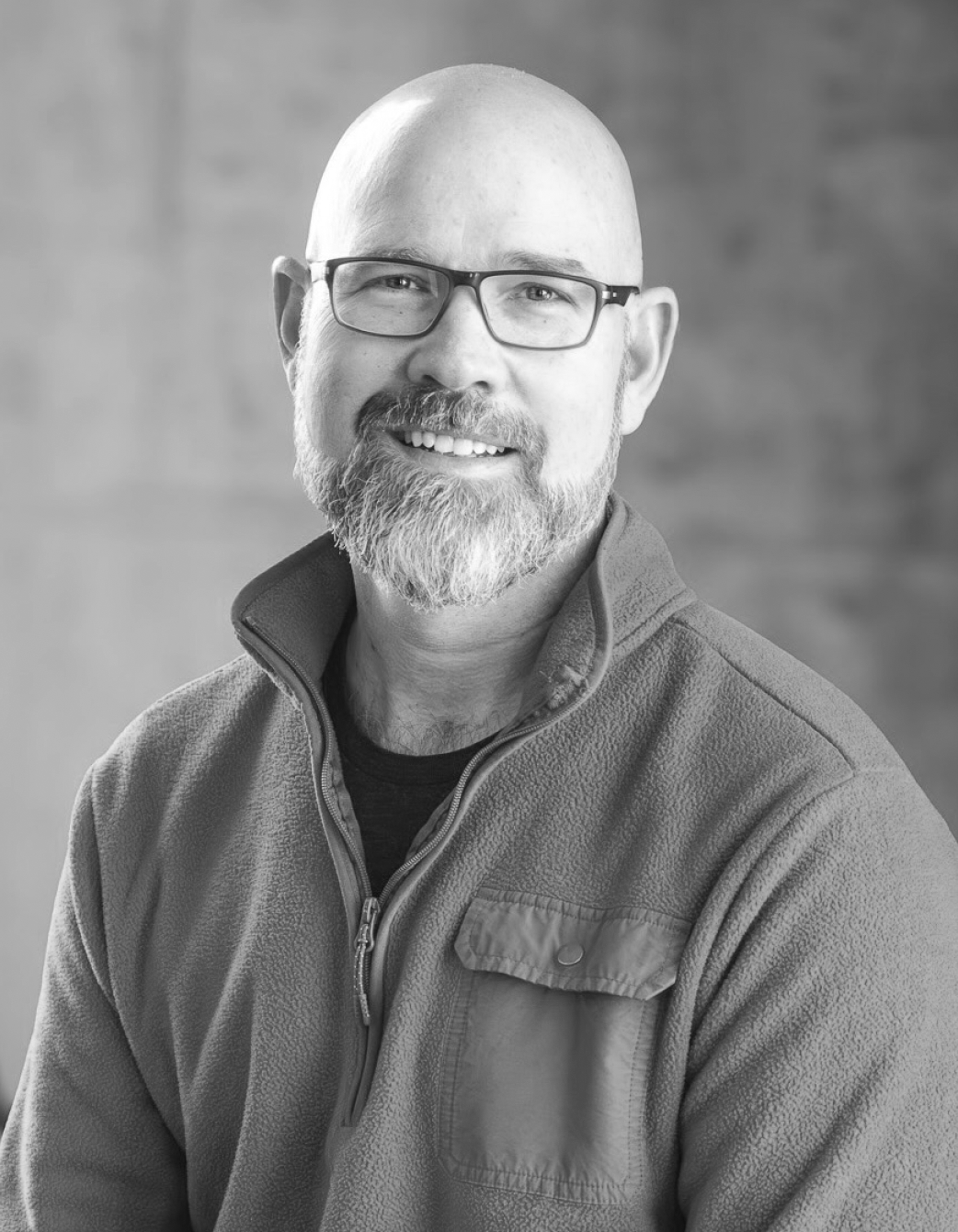 A black and white portrait of a bald man with glasses, a beard, and a mustache, smiling while wearing a fleece jacket.