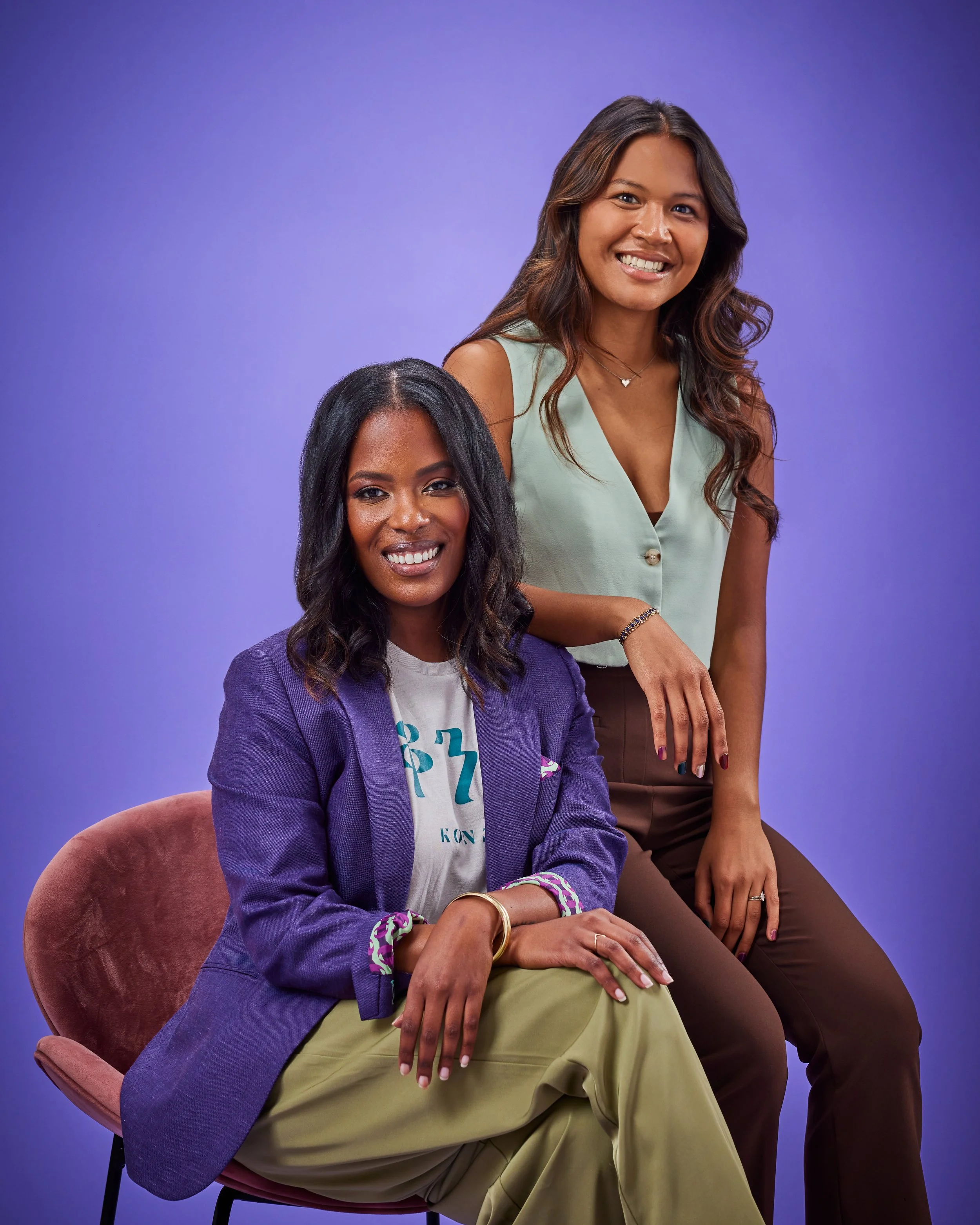 Two women with dark hair smiling, one seated wearing a purple blazer and khaki pants, and one standing wearing a sleeveless light green top and brown pants, against a purple background.