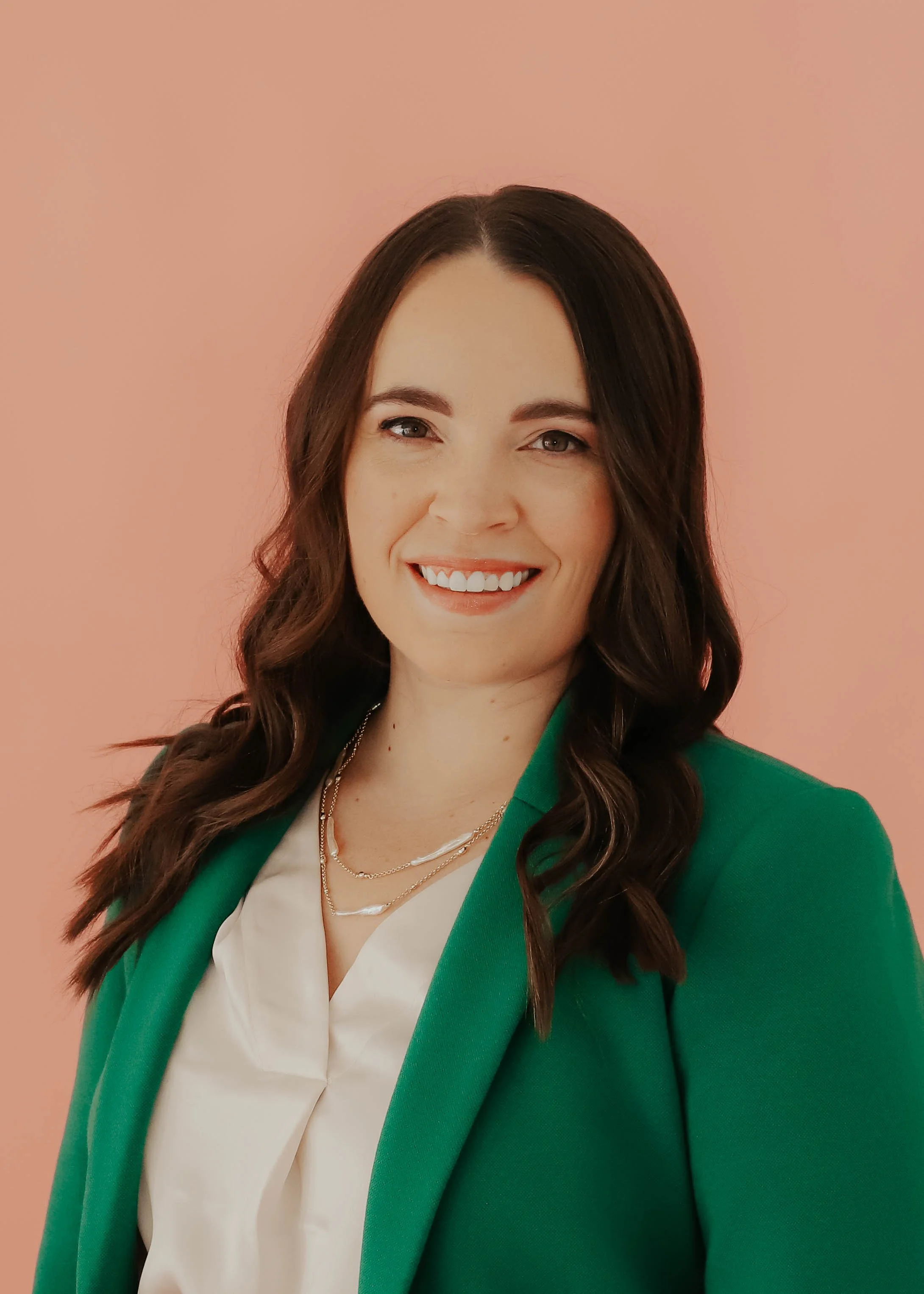 A woman with long, wavy dark brown hair, wearing a green blazer over a white blouse, smiling against a pink background.