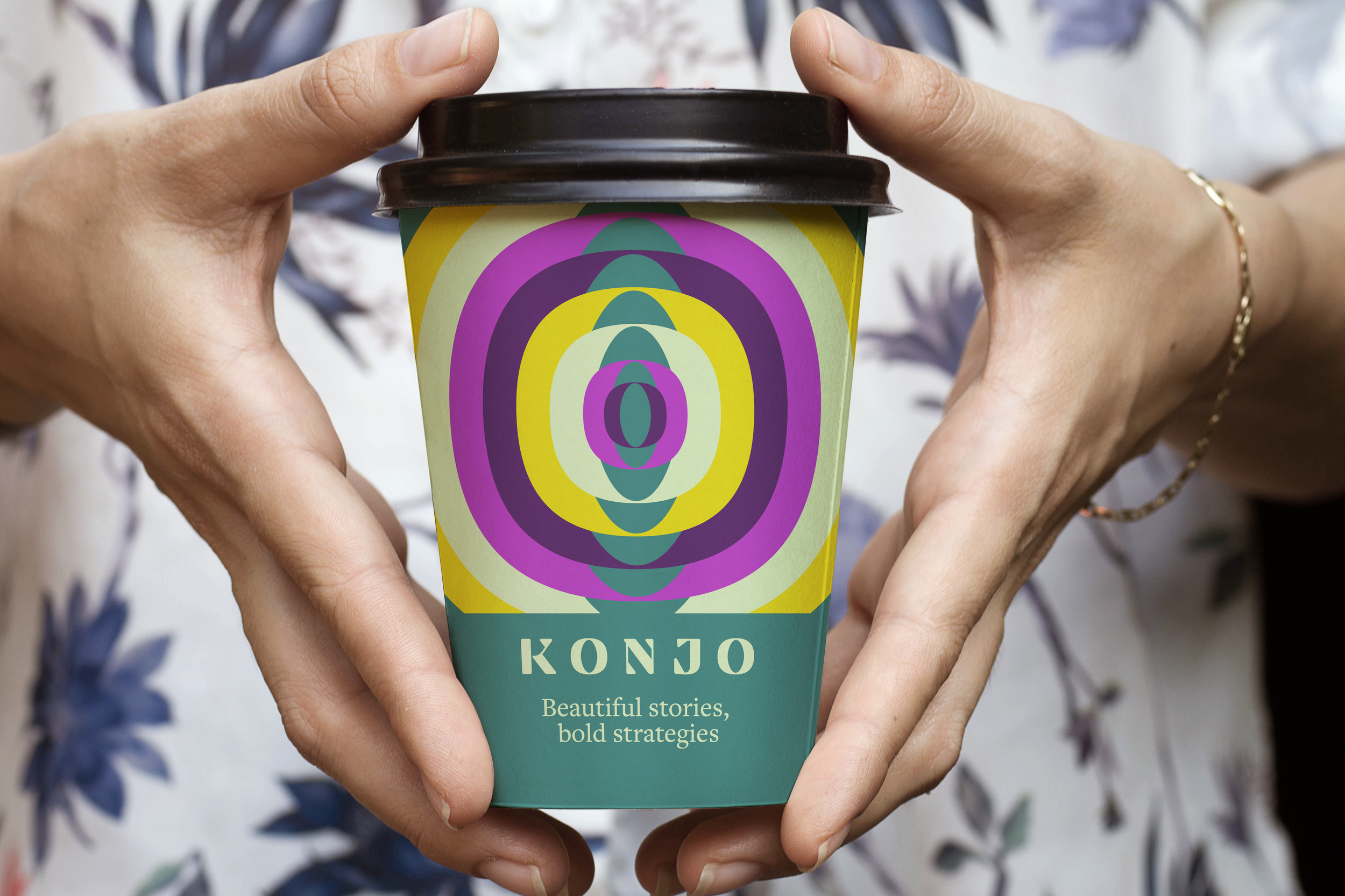 Person holding a colorful cup with a black lid. The cup has a vibrant, abstract, concentric oval pattern in purple, yellow, green, and teal. The label on the cup reads 'KONJO' and 'Beautiful stories, bold strategies.' The person is wearing a floral-patterned shirt and a gold bracelet.