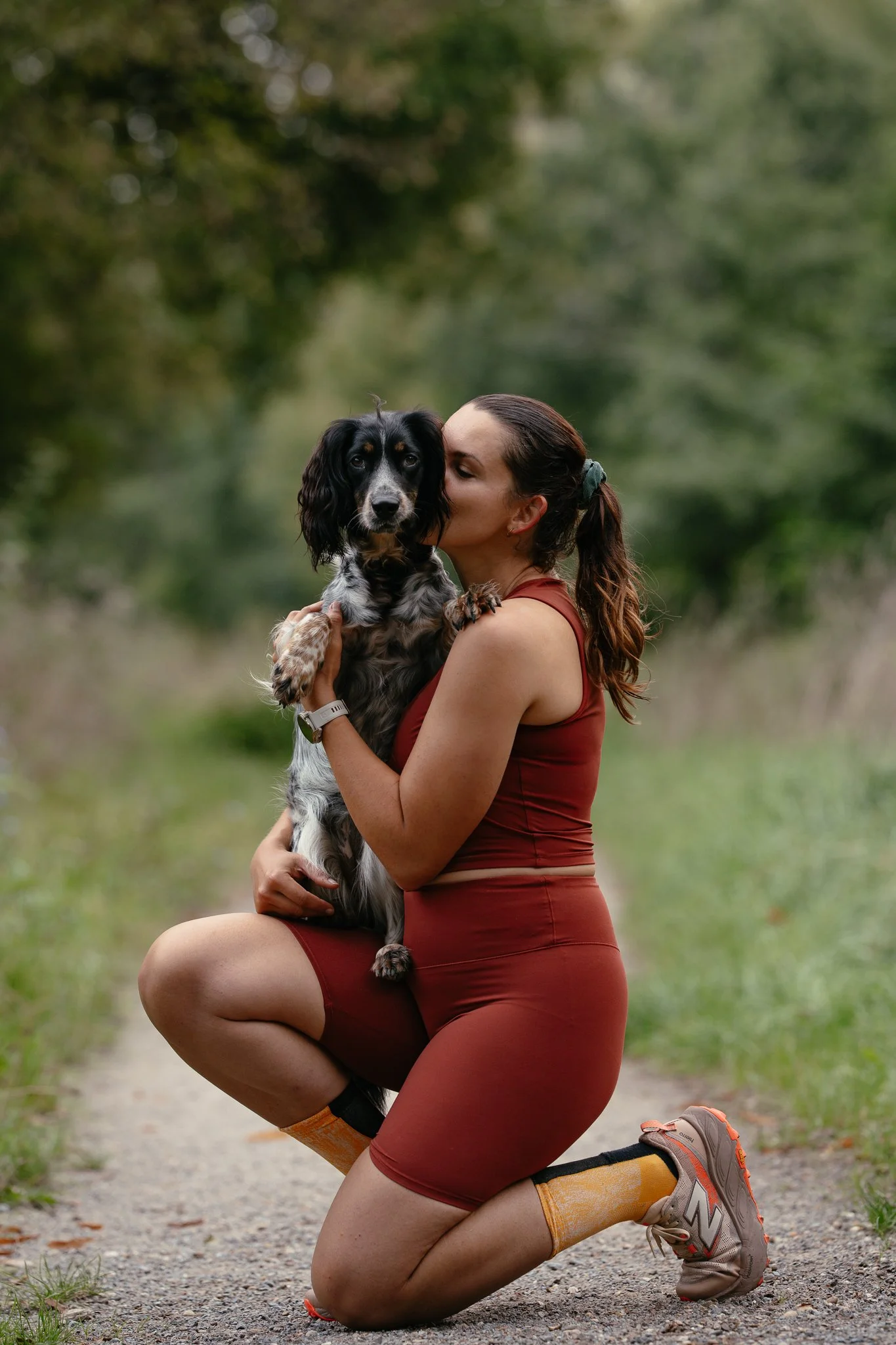 Frau im Sportoutfit kniet im Wald, hält einen  Working Cocker Spaniel und küsst ihn auf den Kopf.